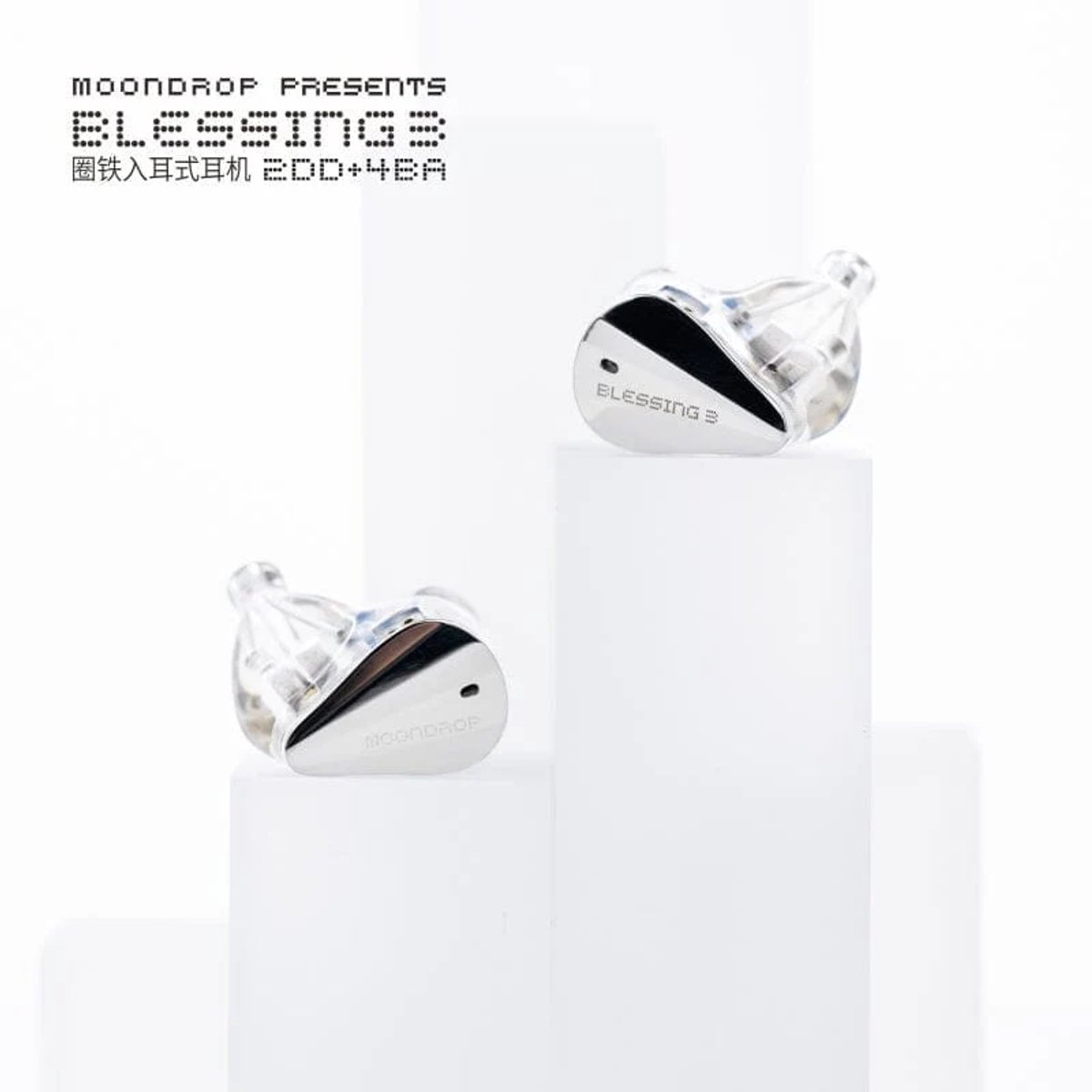 Twee in-ear koptelefoons op witte blokken. Transparant plastic met zilveren accenten. Bovenop staat "Blessing 3". Achtergrond is wit.