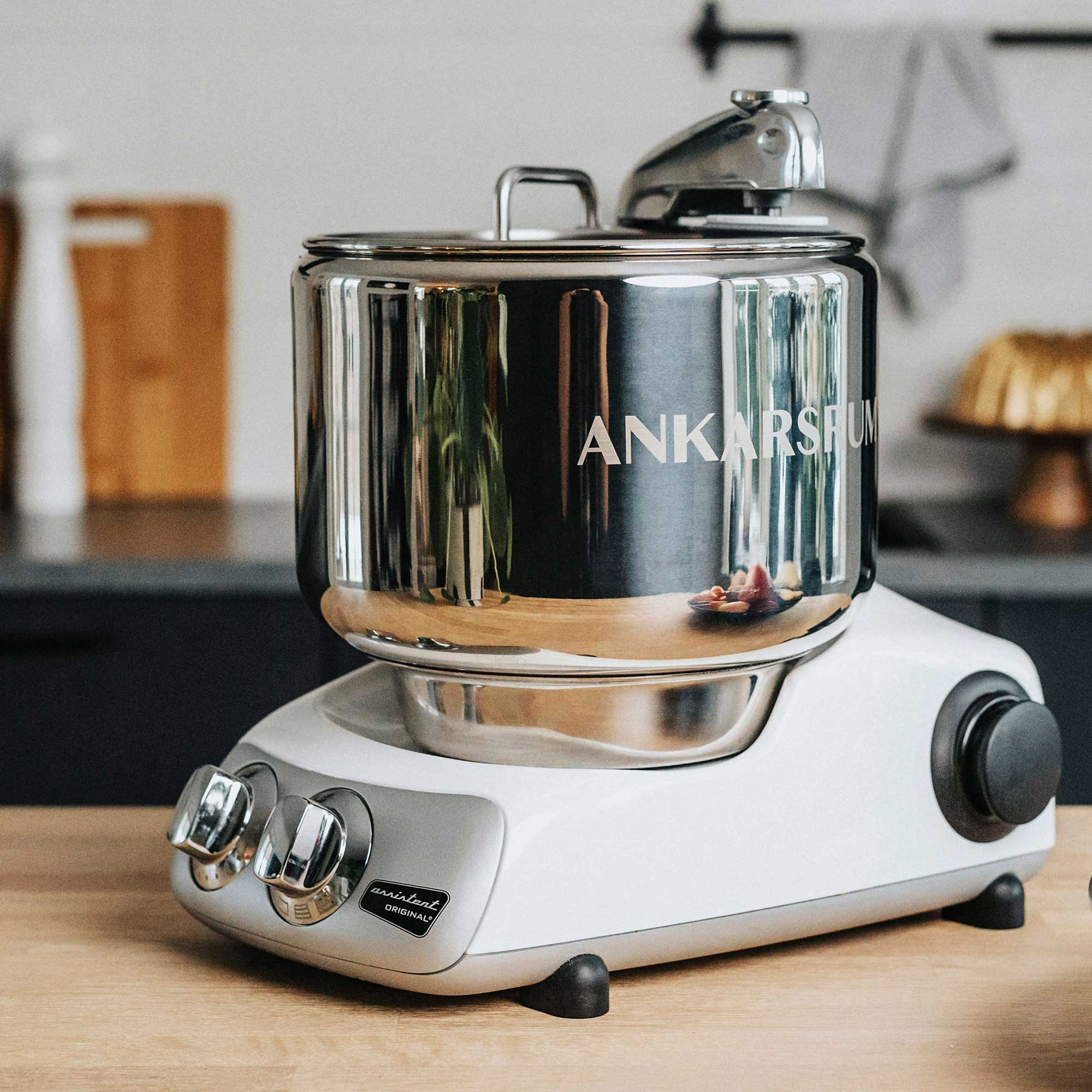 Ein weißer und silberner Ankarsrum-Küchenmixer steht auf einer Holztheke. Eine Edelstahlschüssel und ein Deckel sind darauf.