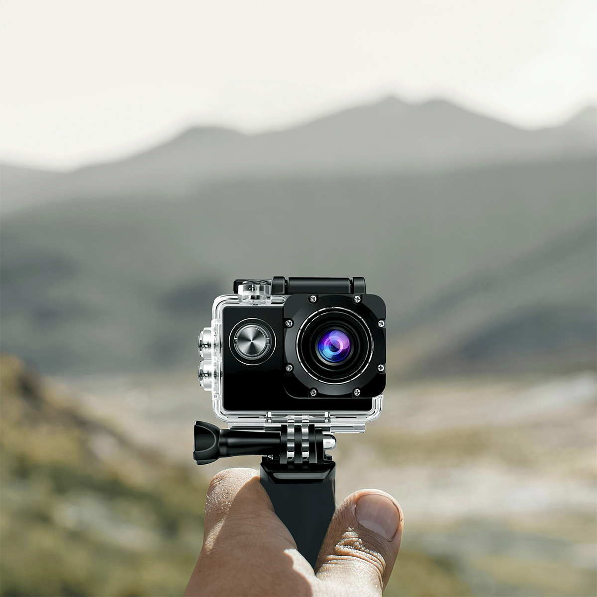 Ręka trzyma czarną kamerę sportową z przezroczystą obudową. Rozmyte góry w tle.