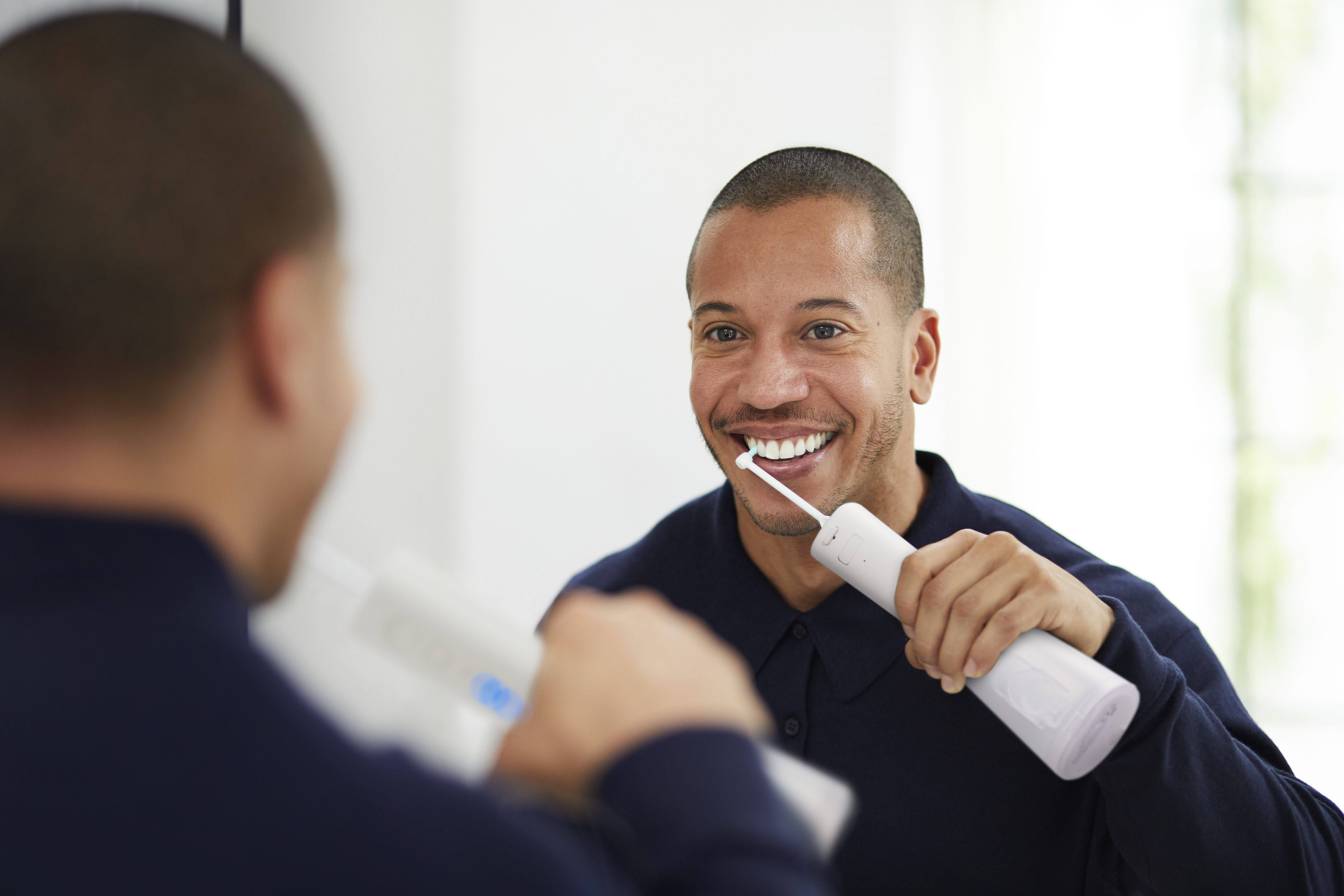 Mężczyzna myje zęby irygatorem przed lustrem. Uśmiecha się. Odbija się inny mężczyzna.