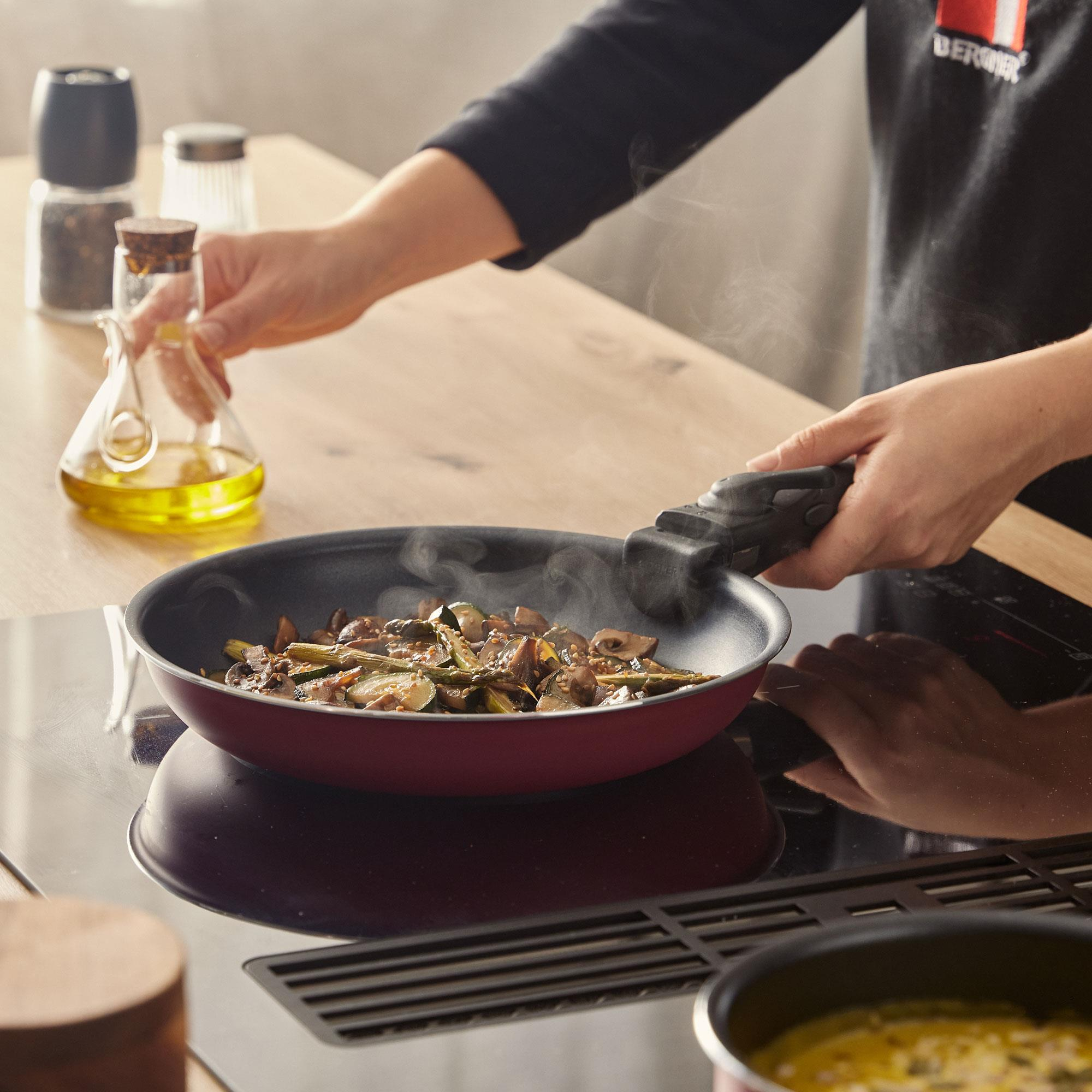 Una persona cocina verduras en una sartén. Botella de aceite en la mano, sale vapor. Escena de cocina con mesa de madera y estufa.