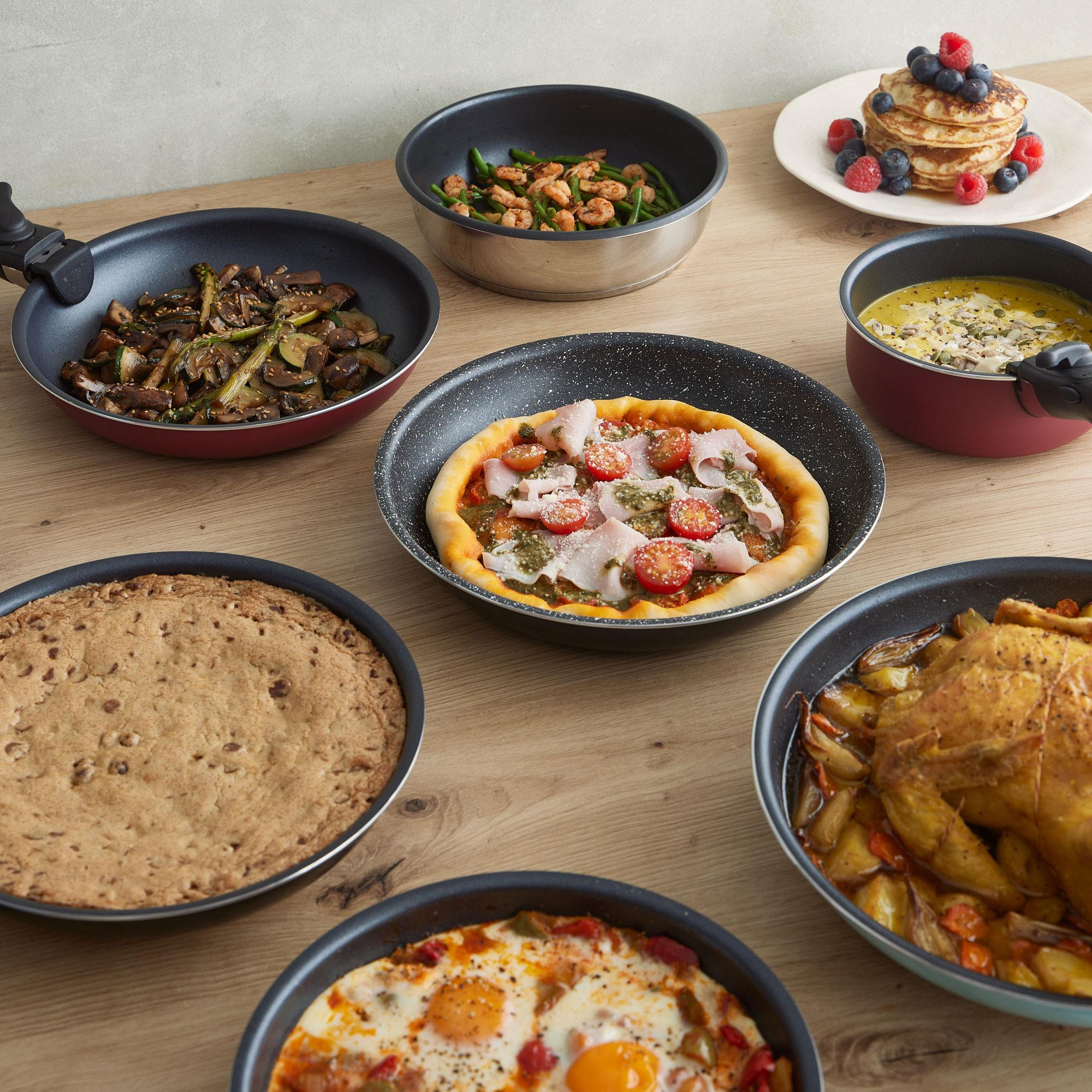 Mesa de madera con varios platos: pizza, panqueques, verduras, sopa, pollo asado y huevos.