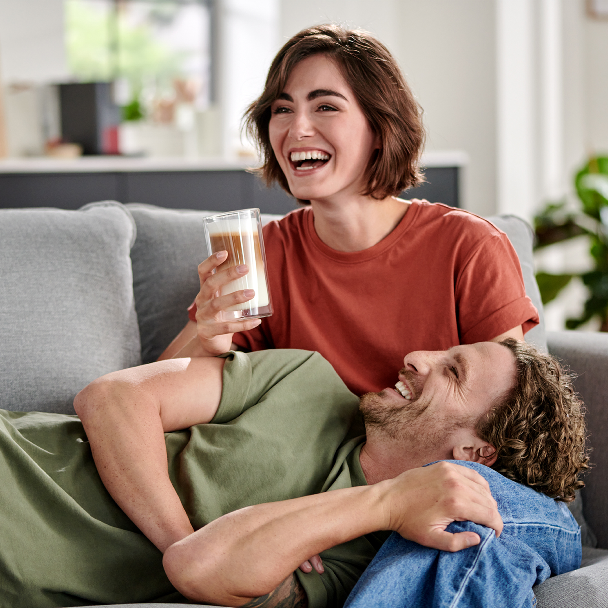 Ein lachendes Paar auf dem Sofa. Frau hält Getränk. Mann liegt auf ihrem Schoß. Wohnraum sichtbar.