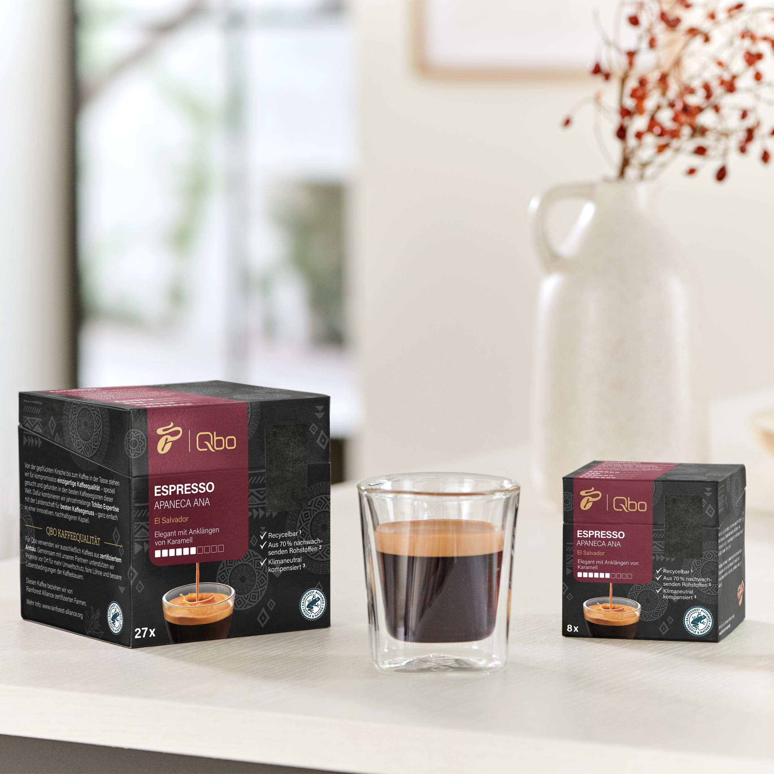 Espresso-Tasse neben zwei Kaffeeboxen. Eine Box ist größer und zeigt Kaffeedetails. Eine Vase mit roten Blumen ist im Hintergrund.