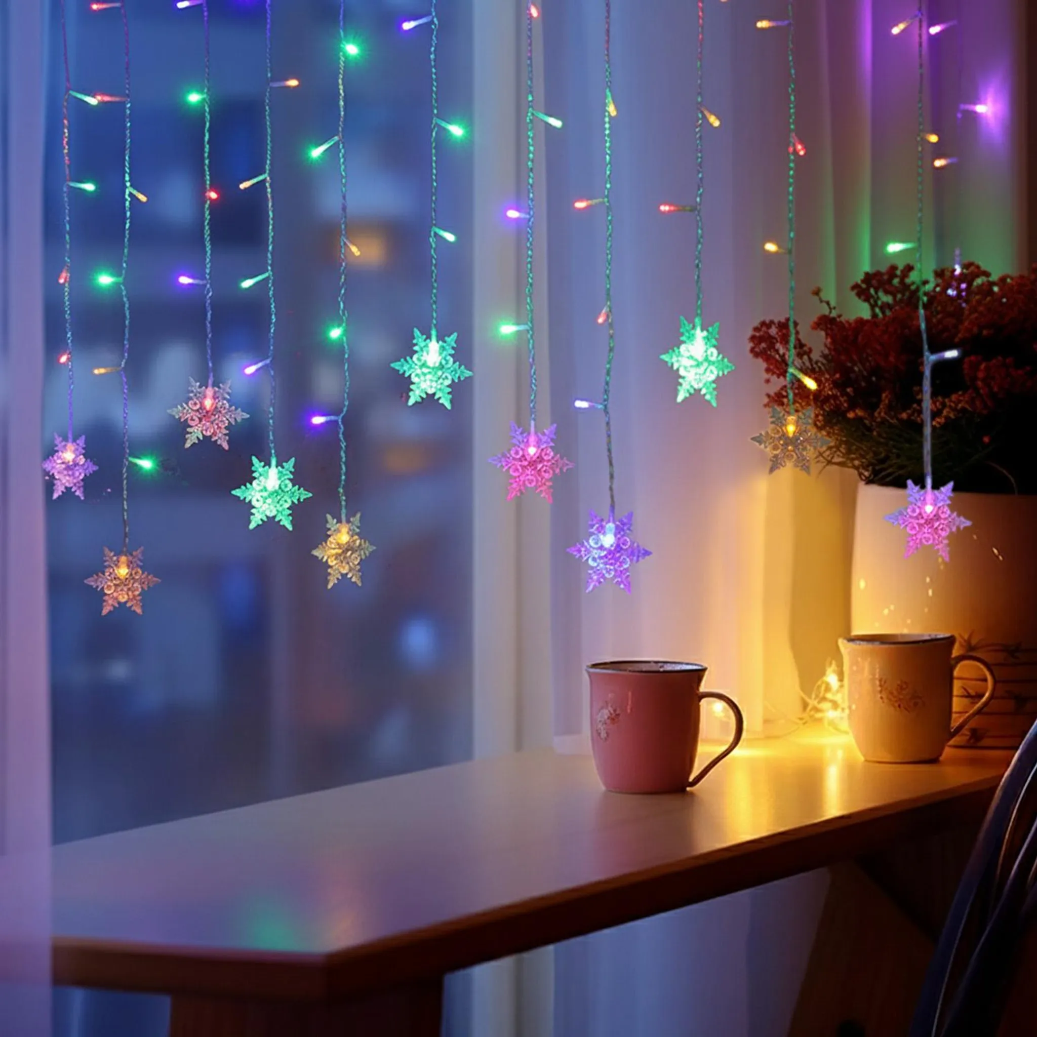 Lichterketten und Schneeflockenlichter hängen in der Nähe eines Fensters mit zwei Tassen auf einer Holzoberfläche. Pflanzen im Hintergrund.