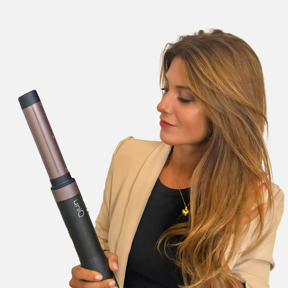 Donna che tiene un arricciacapelli. Indossa una giacca beige e un top nero. I suoi lunghi capelli sono castani. Lo sfondo è bianco.