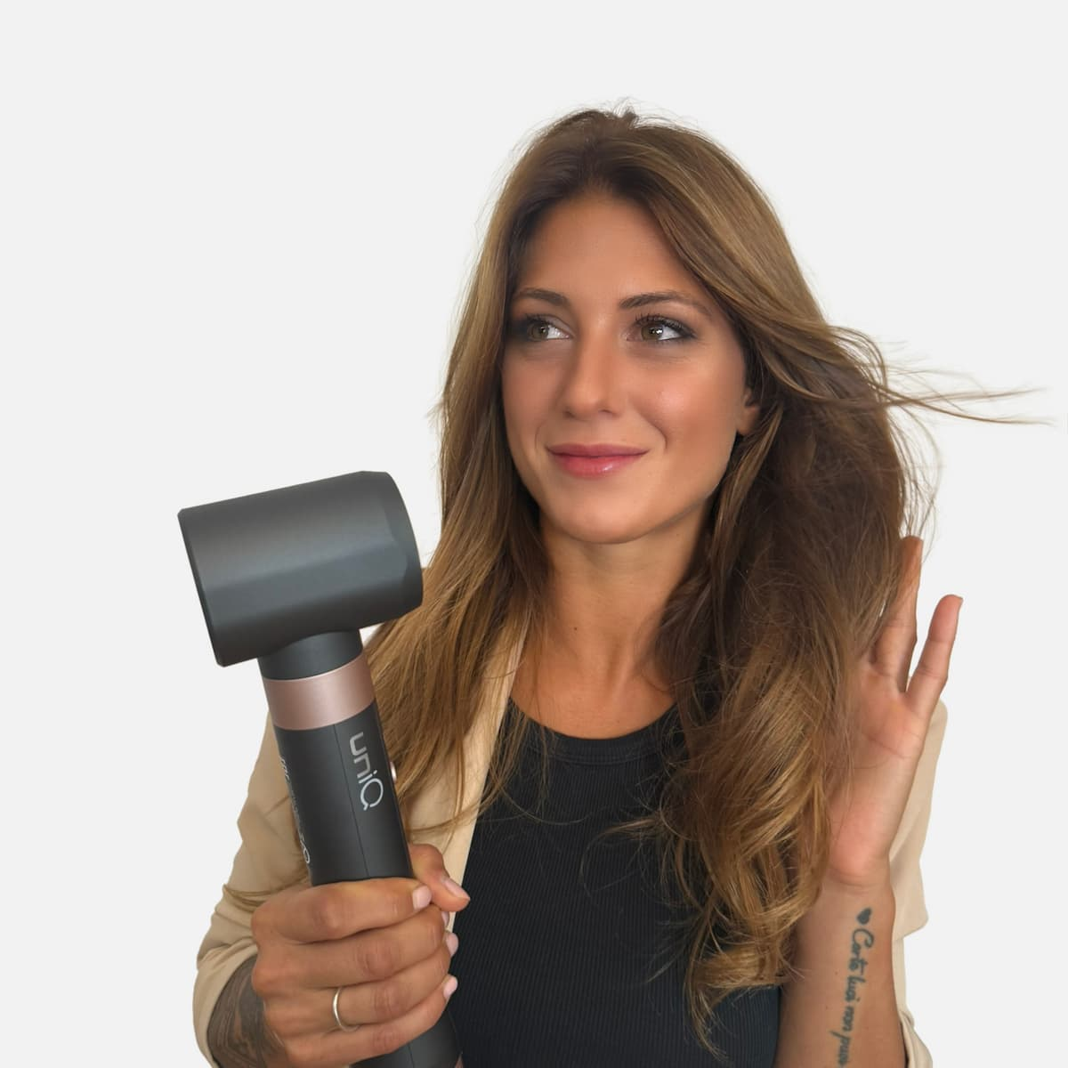 Una donna tiene un asciugacapelli, guardando di lato. Indossa un top nero, un blazer beige e ha lunghi capelli castani.