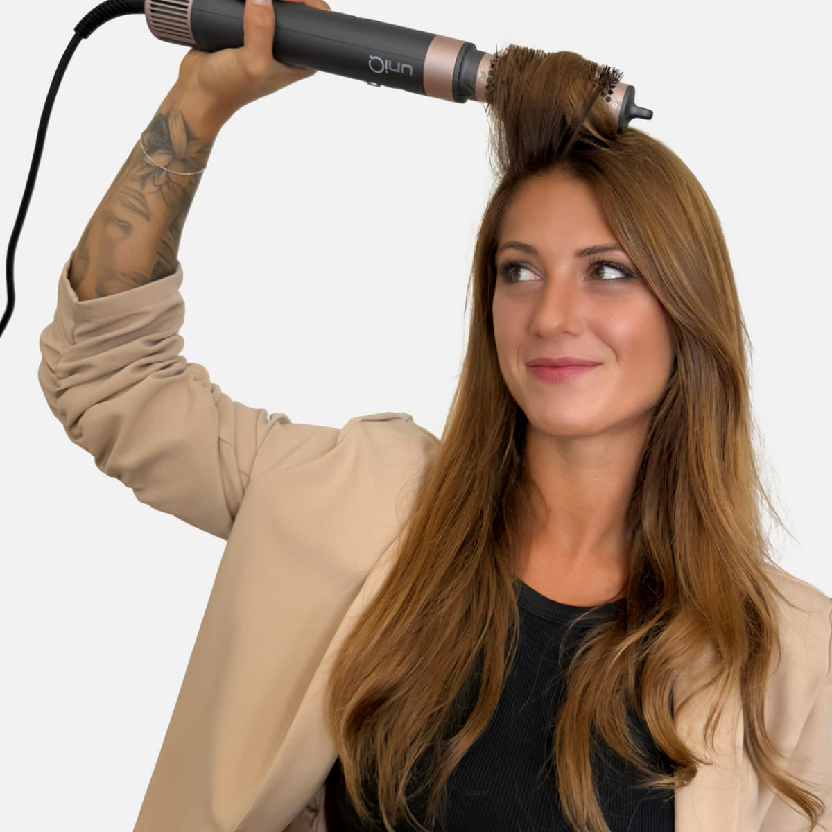 Una donna usa uno strumento per lo styling dei capelli. Tiene un asciugacapelli, guarda di lato, con lunghi capelli castani e una giacca beige.