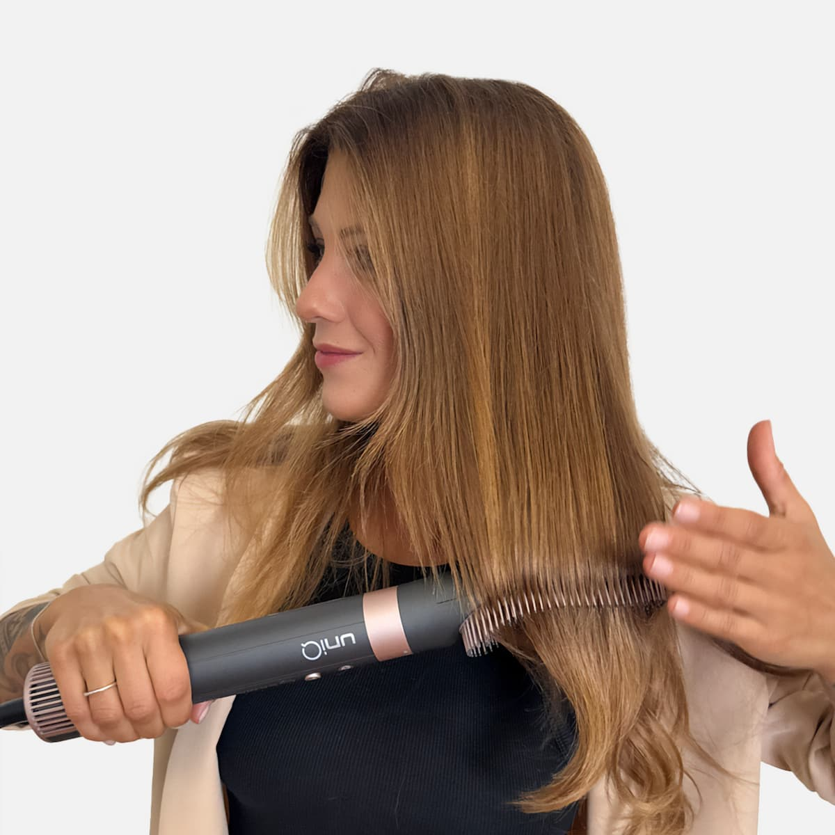 Una donna con i capelli biondi usa uno strumento per lo styling dei capelli. Indossa una giacca beige e un top nero. Lo sfondo è bianco.