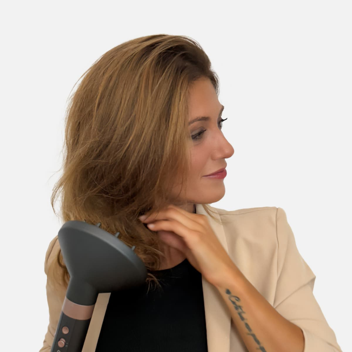 Una donna con capelli castano ondulati, tiene un diffusore per capelli. Indossa un top nero e un blazer beige, guardando di lato. Un tatuaggio visibile.