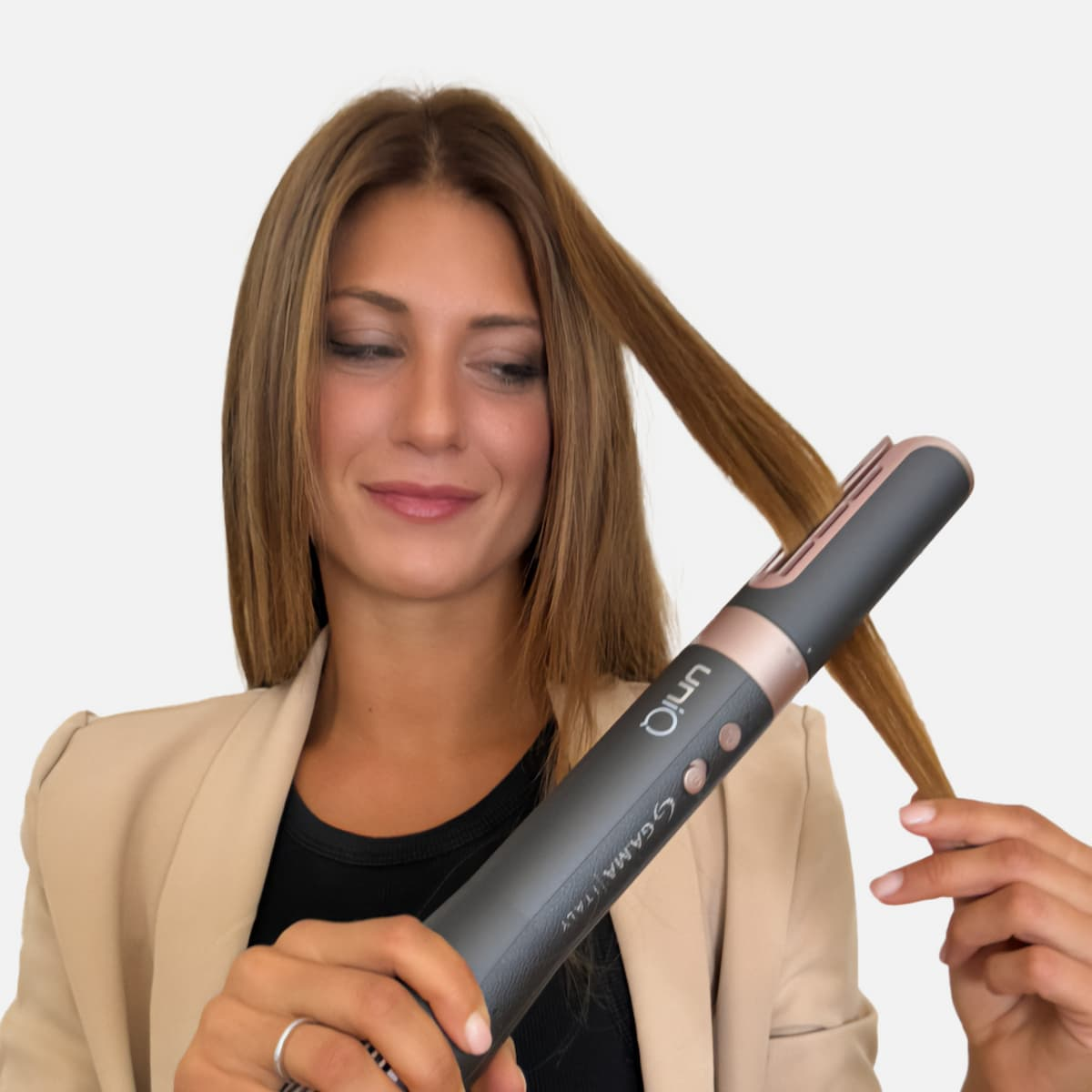 Una donna che tiene in mano una piastra per capelli, guardando i suoi capelli. Indossa una giacca beige e una camicia nera. Lo sfondo è bianco.