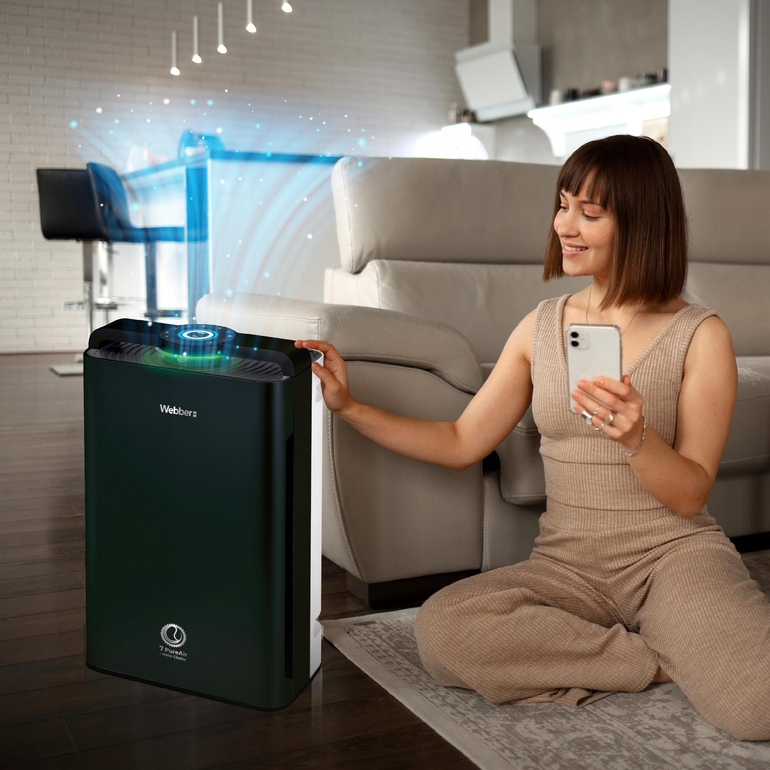 Kobieta siedzi w pobliżu oczyszczacza powietrza, trzymając telefon. Uśmiecha się. Za nią jest kanapa.