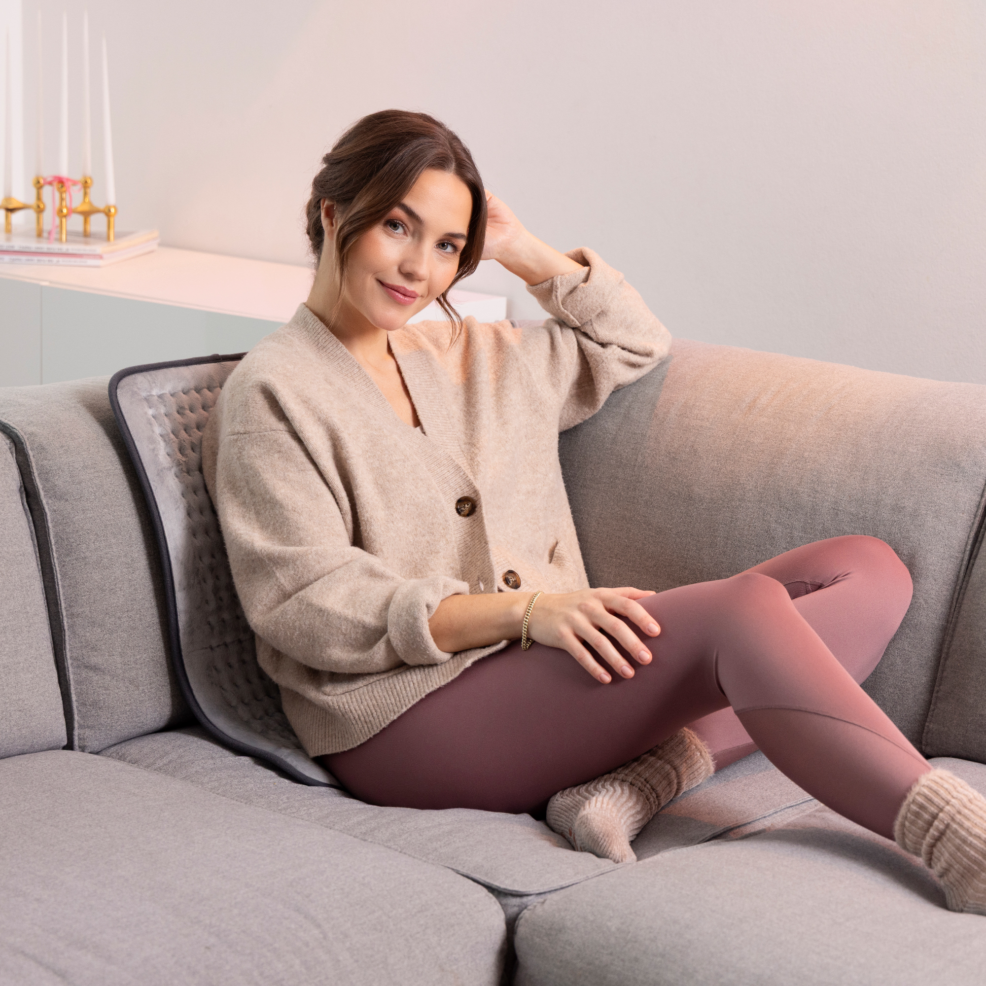 Mujer con cárdigan y leggings beige sentada en un sofá gris con una almohadilla térmica. Sonríe a la cámara.