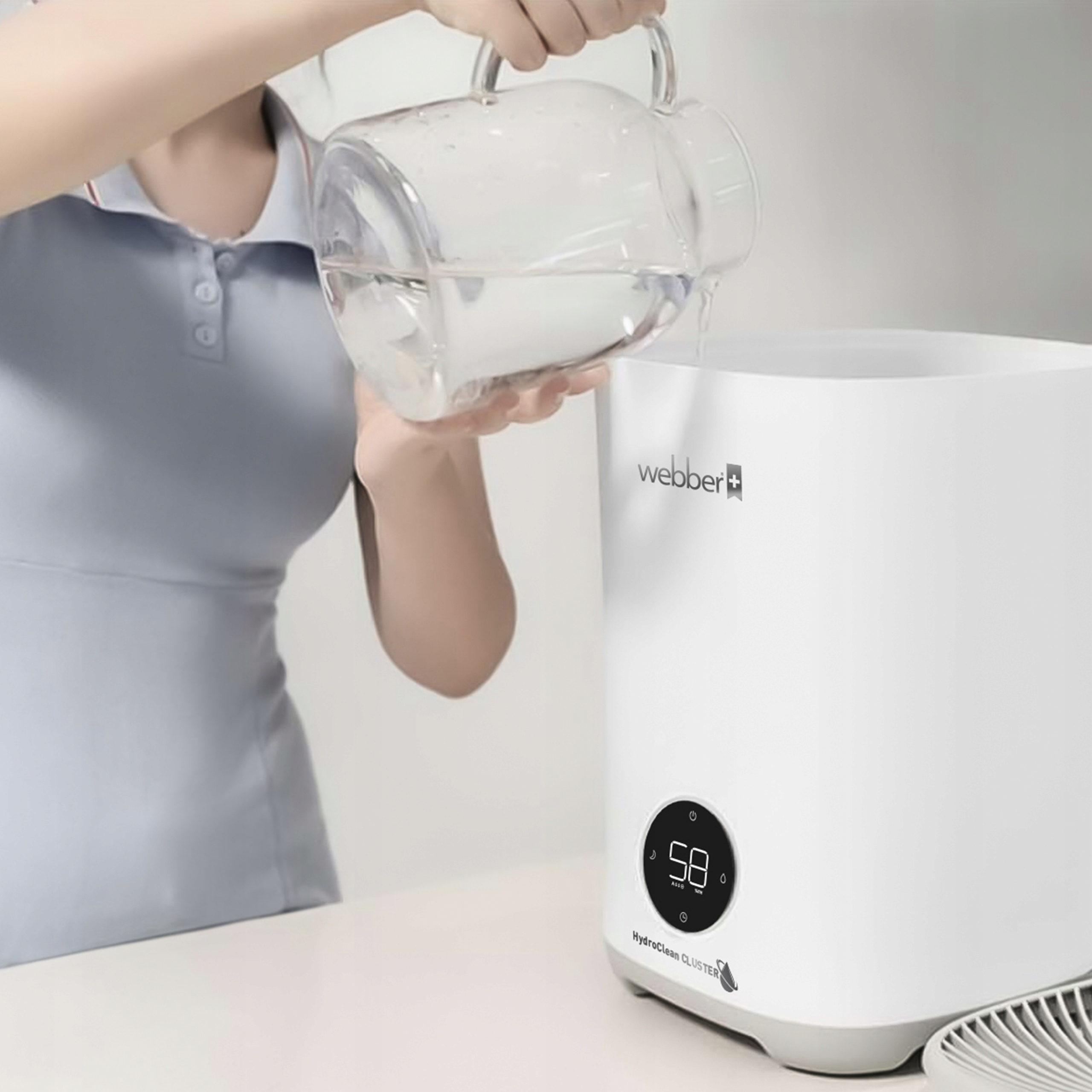 Osoba wlewa wodę z dzbanka do białego oczyszczacza powietrza. Oczyszczacz wyświetla 58 i HydroClean CLUSTER.