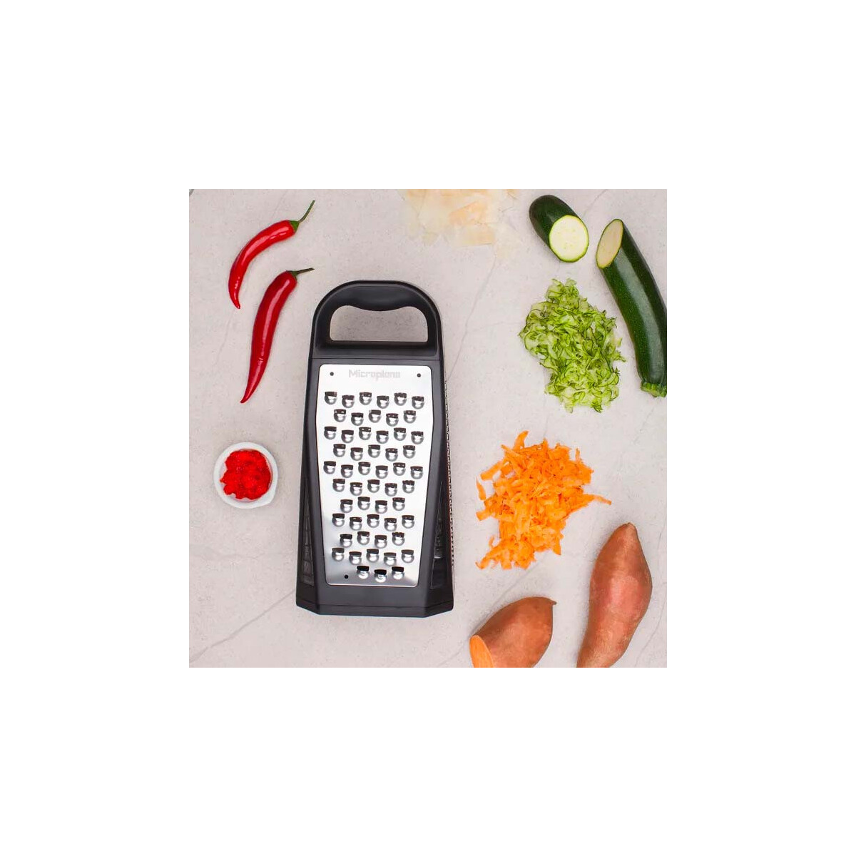 Eine Reibe, rote Paprika, geriebenes Gemüse, Zucchini und Süßkartoffeln sind auf einer hellen Oberfläche angeordnet.