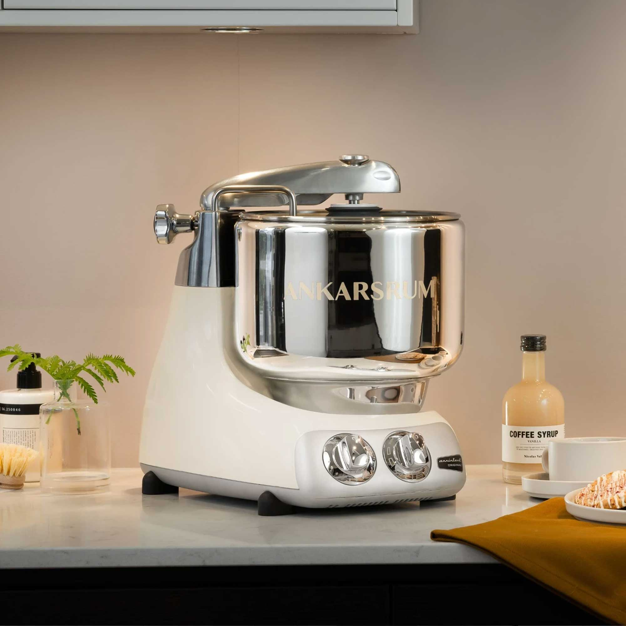Ein cremefarbener Ankarsrum-Küchenmixer mit Chromakzenten auf einer Arbeitsplatte. Kaffeesirup und Gebäck sind sichtbar.