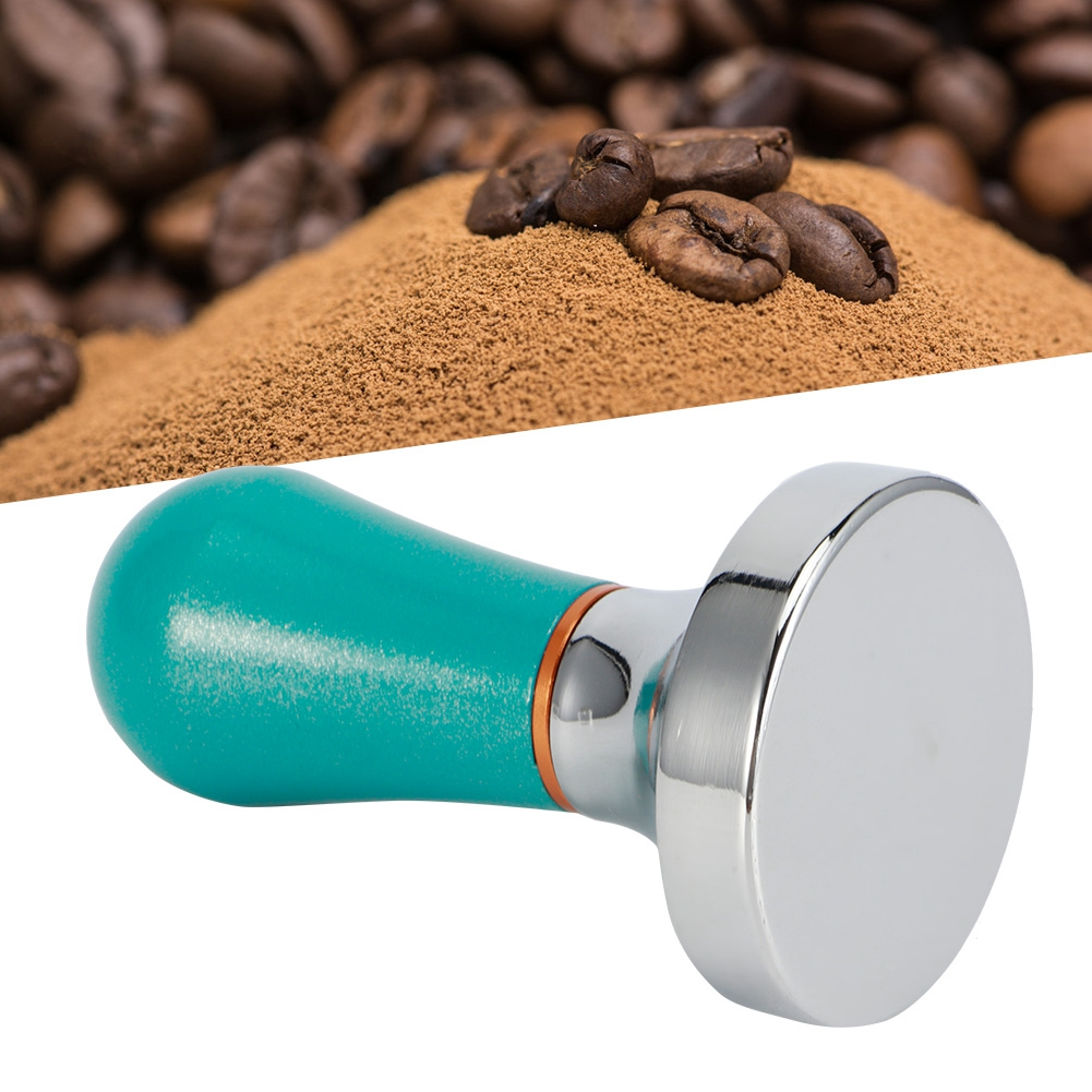 Kaffeebohnen und gemahlener Kaffee im Hintergrund. Ein Tamper mit tealfarbenem Griff und silberner Basis im Vordergrund.