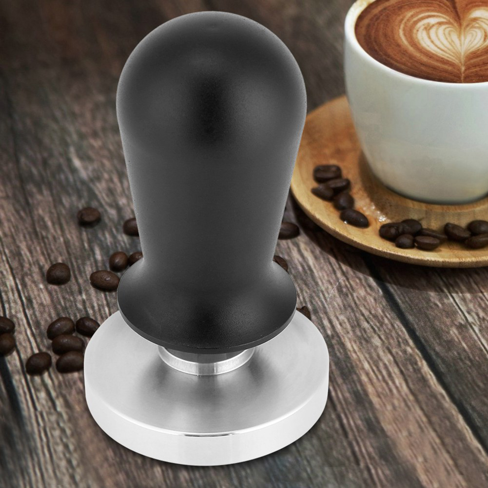 Ein Kaffeestempel mit schwarzem Griff und silberner Basis. Kaffeebohnen liegen auf einer Holzoberfläche, mit einer Kaffeetasse.