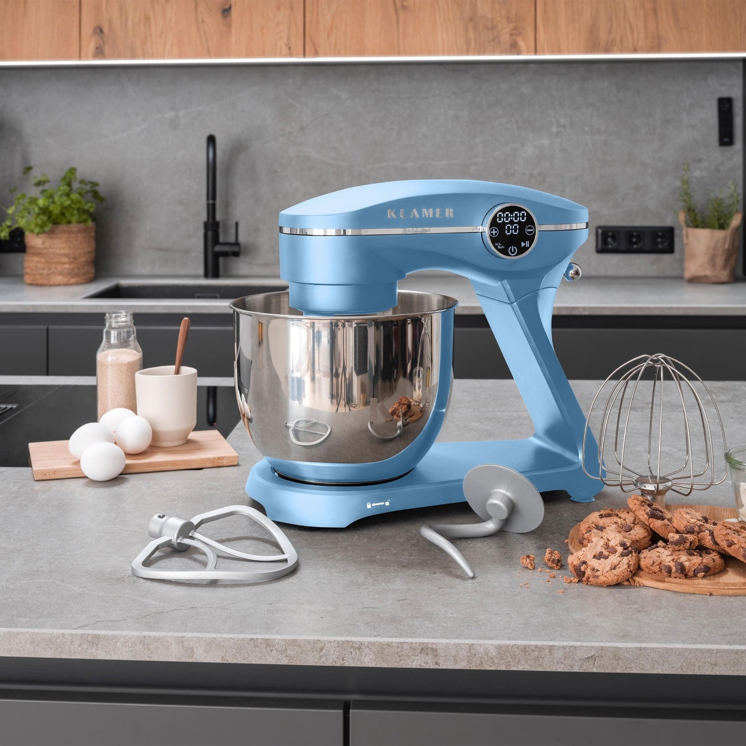 Ein blauer Küchenmixer steht auf einer Arbeitsplatte mit Zutaten. Kekse und ein Schneebesen befinden sich neben dem Mixer. Eier und ein Glas sind in der Nähe.