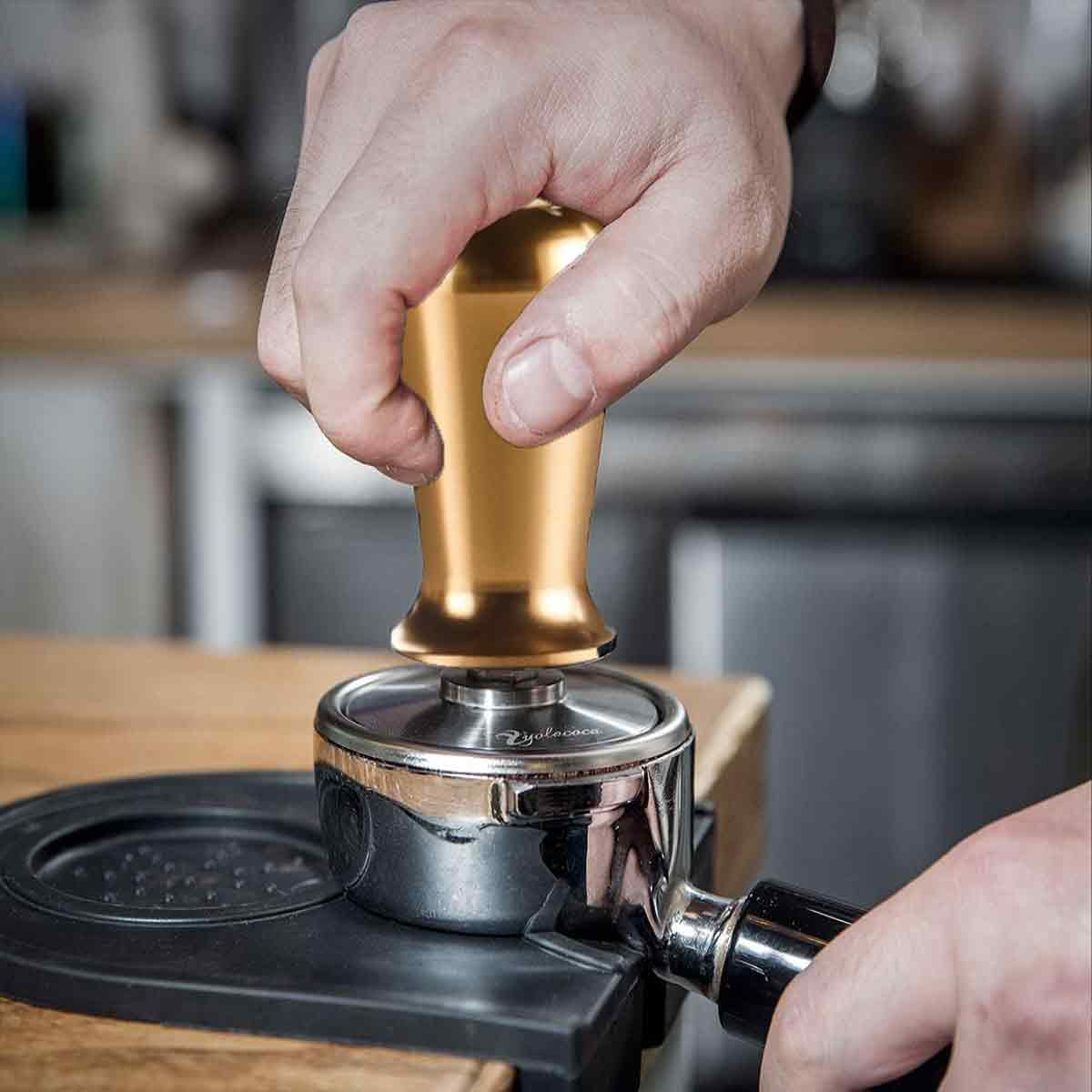 Eine Hand drückt einen goldenen Tamper auf einen Siebträger. Der Siebträger befindet sich auf einer schwarzen Matte. Der Hintergrund ist eine verschwommene Küche.