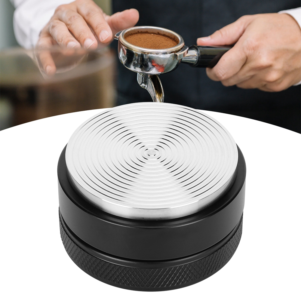 Ein Kaffeetamper steht auf einem Tisch. Oben hält eine Person einen Siebträger mit Kaffeepulver. Der Tamper ist schwarz und silber.