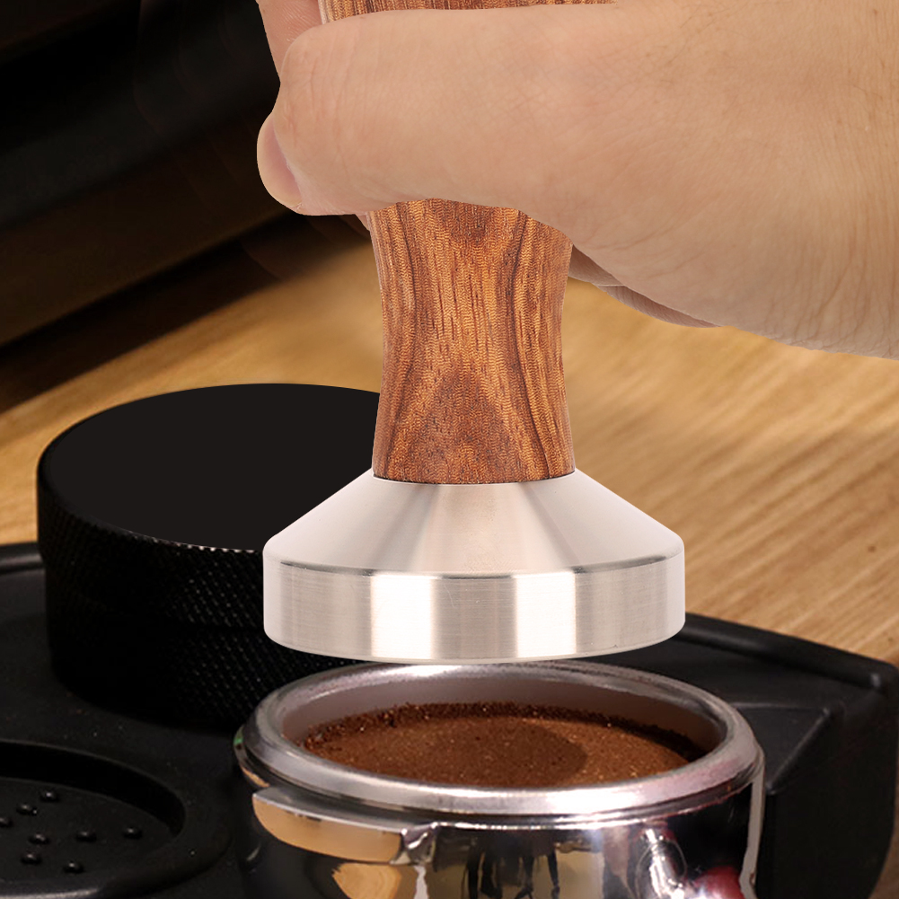 Eine Hand drückt einen Tamper mit Holzgriff auf Kaffeepulver in einem Siebträger. Im Hintergrund befindet sich ein schwarzes Objekt.