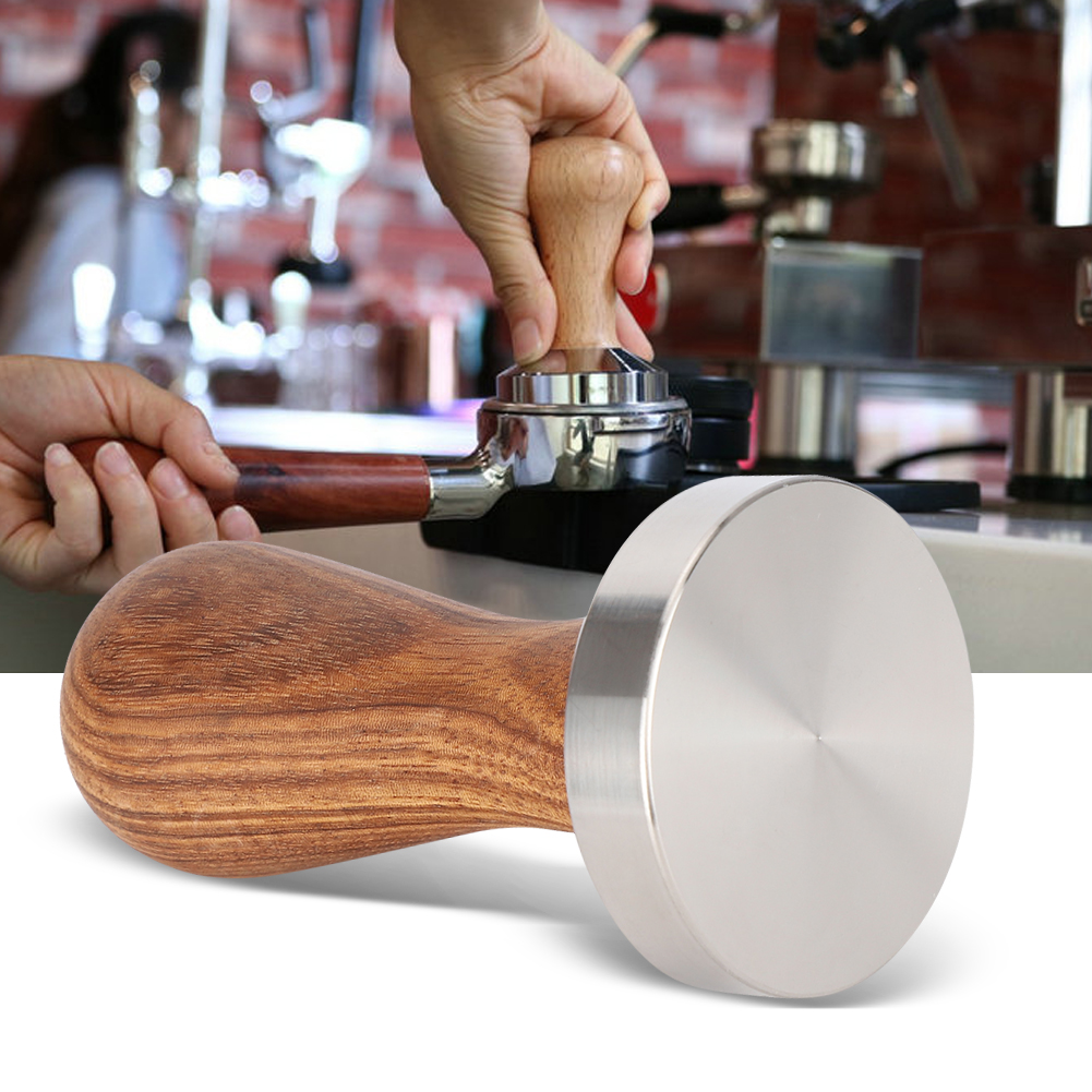 Ein Barista presst Kaffeepulver in einem Siebträger mit einem Tamper. Der Tamper hat einen Holzgriff und einen Metallsockel. Im Hintergrund steht eine Kaffeemaschine.