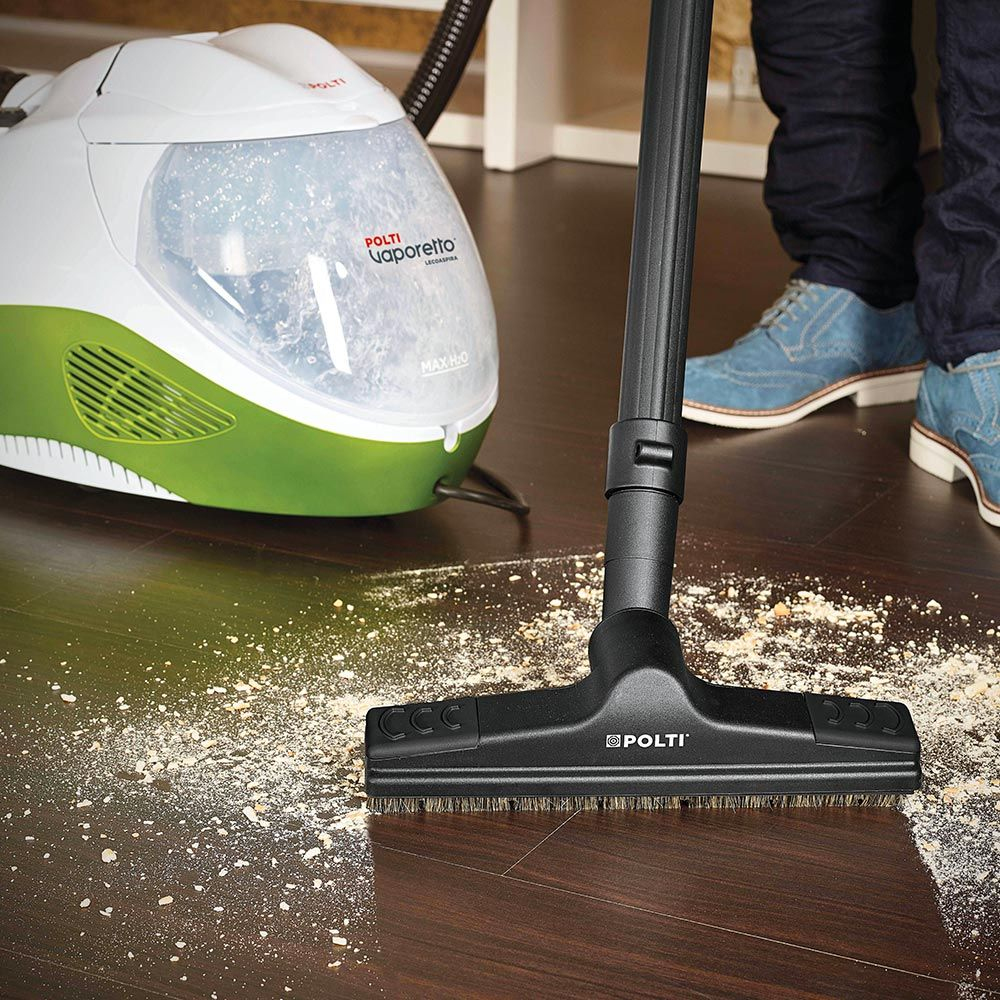 Una persona usa una limpiadora de vapor Polti en un piso de madera, quitando escombros. La máquina es verde y blanca. La persona lleva vaqueros y zapatos azules.