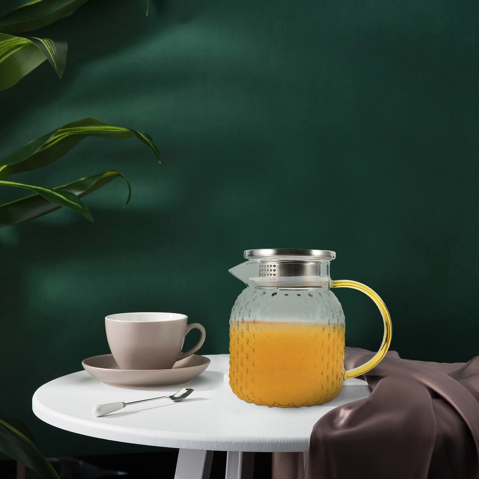 Ein Tisch mit einer Teetasse, einem Löffel und einer Glaskanne mit orangefarbener Flüssigkeit. Grüner Hintergrund und eine Pflanze.