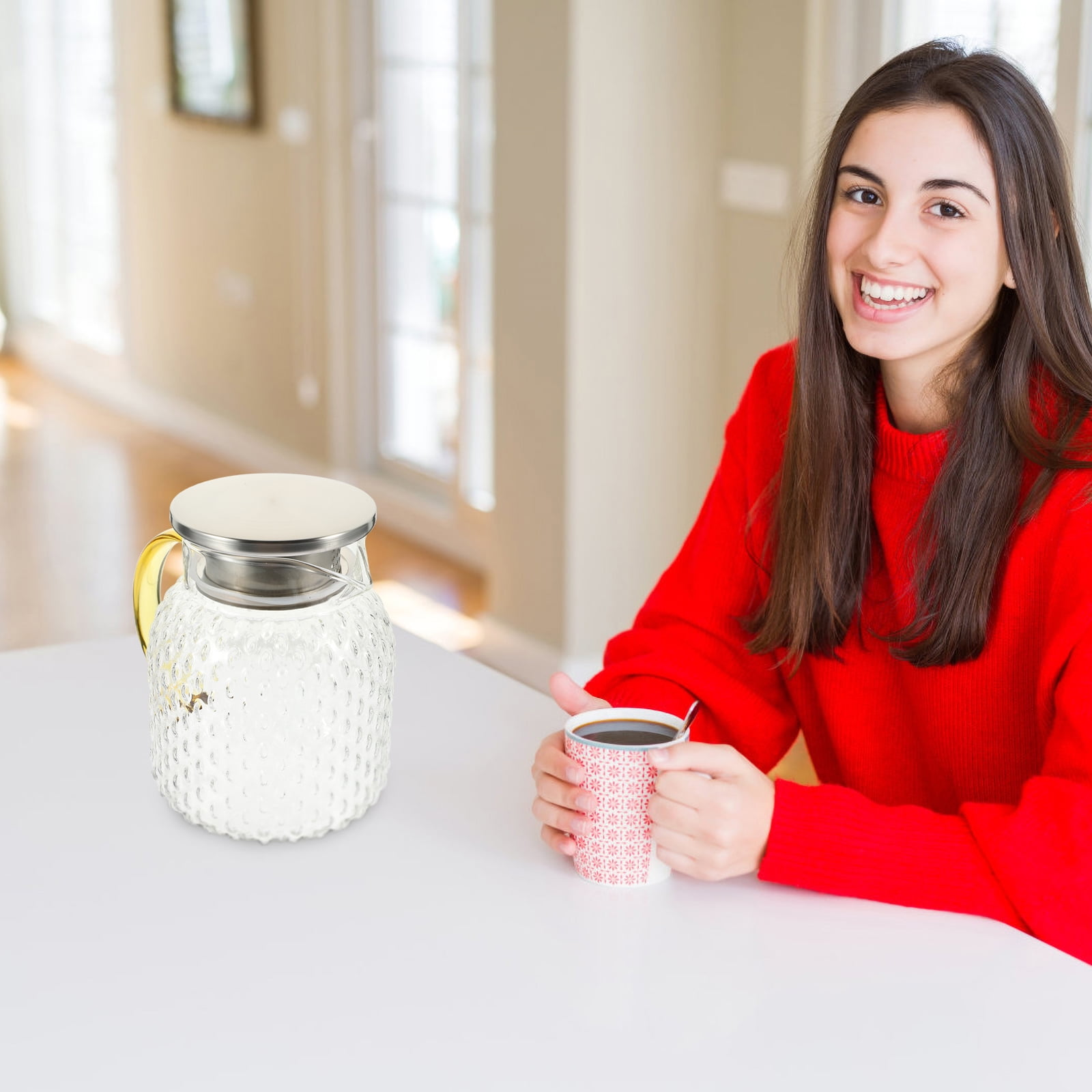 Eine Frau in einem roten Pullover lächelt und hält eine Kaffeetasse. Auf dem Tisch steht eine Glaskanne mit Milch.