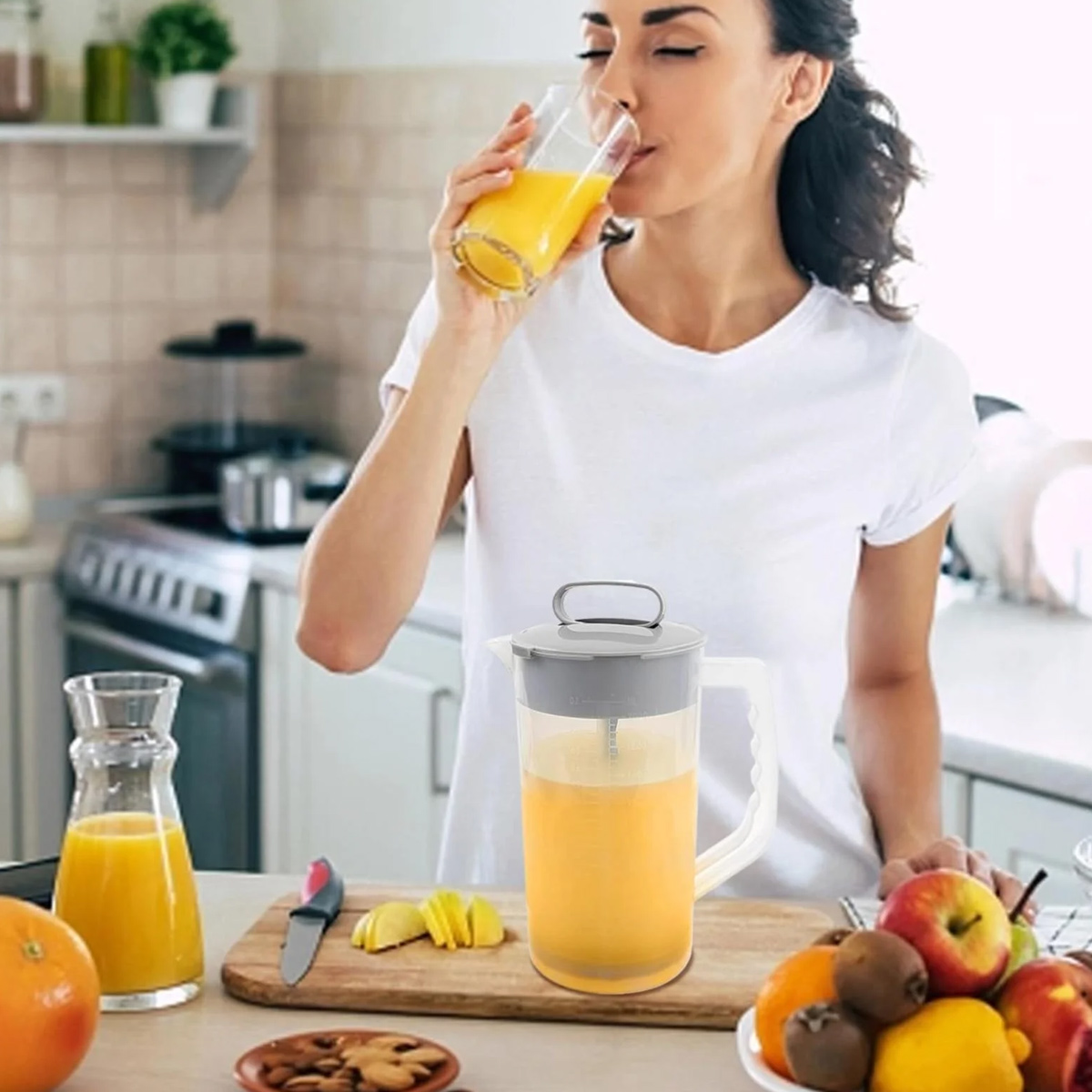 Eine Frau trinkt Orangensaft in einer Küche. Ein Entsafter, Früchte und ein Schneidebrett sind im Vordergrund.
