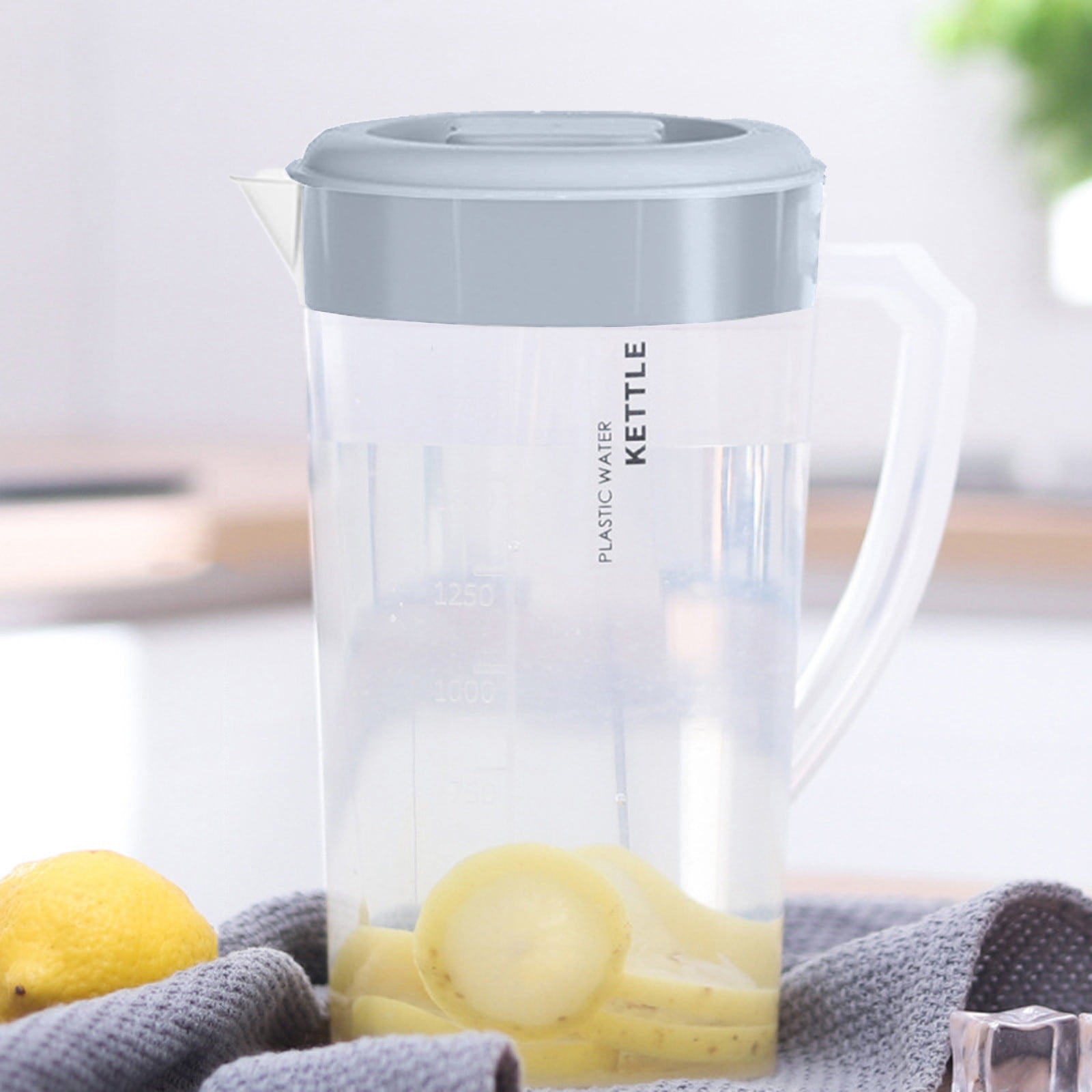 Ein klarer Wasserkessel aus Kunststoff mit Wasser und Obstscheiben. Eine Zitrone und ein graues Handtuch im Vordergrund.