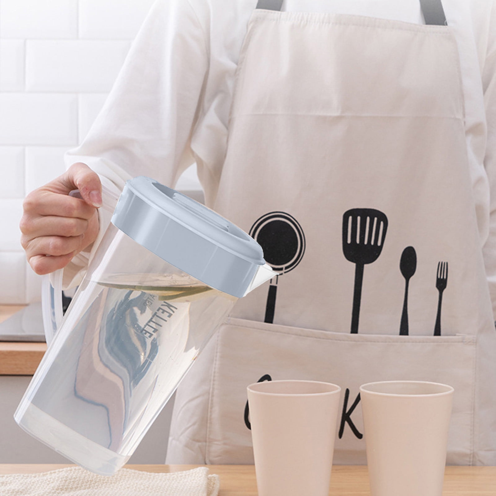 Eine Person gießt Wasser aus einem Krug in eine Tasse. Schürze mit Küchenutensilien und zwei Tassen.