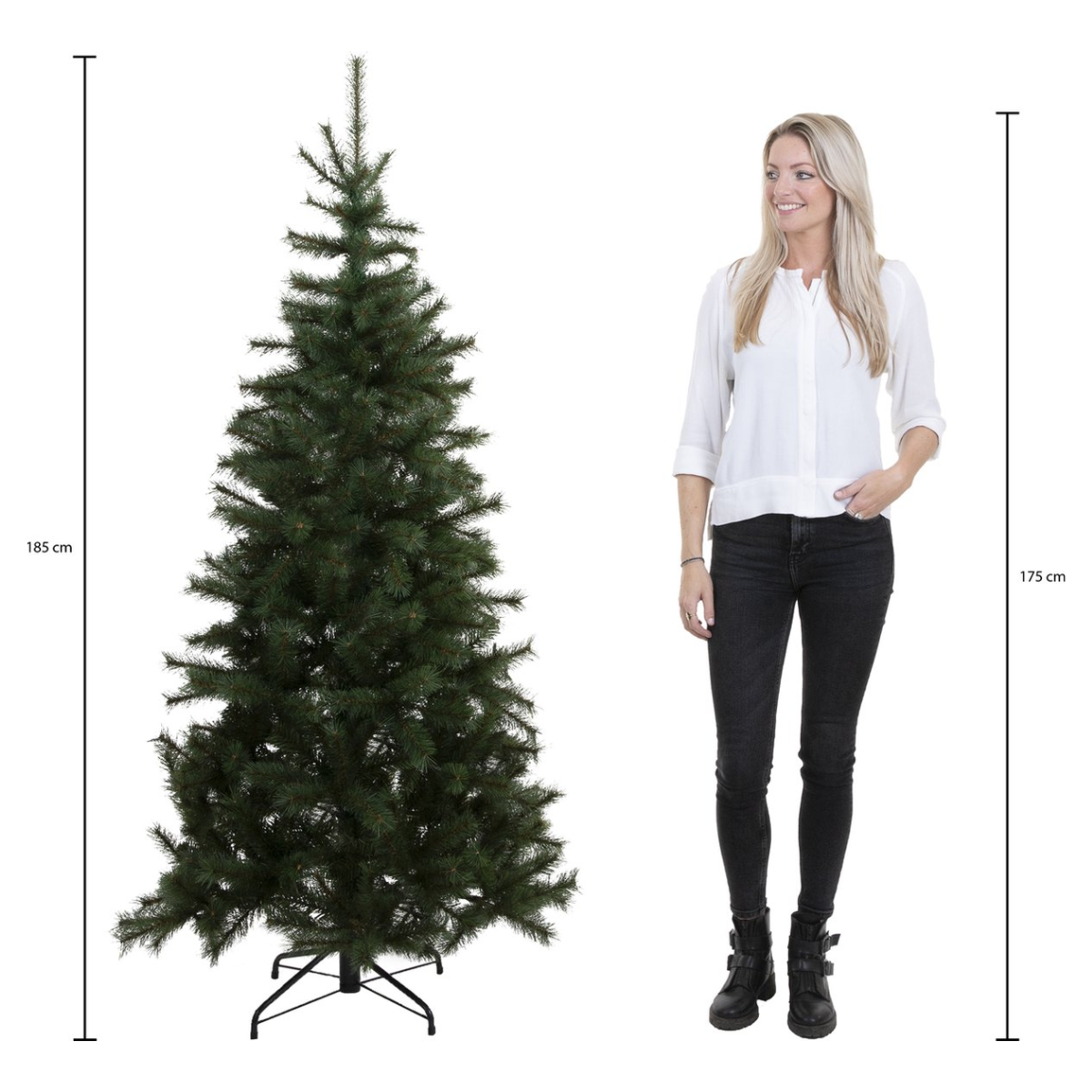Une femme se tient à côté d'un grand sapin de Noël artificiel. L'arbre est vert foncé. La femme porte une chemise blanche et un jean noir.