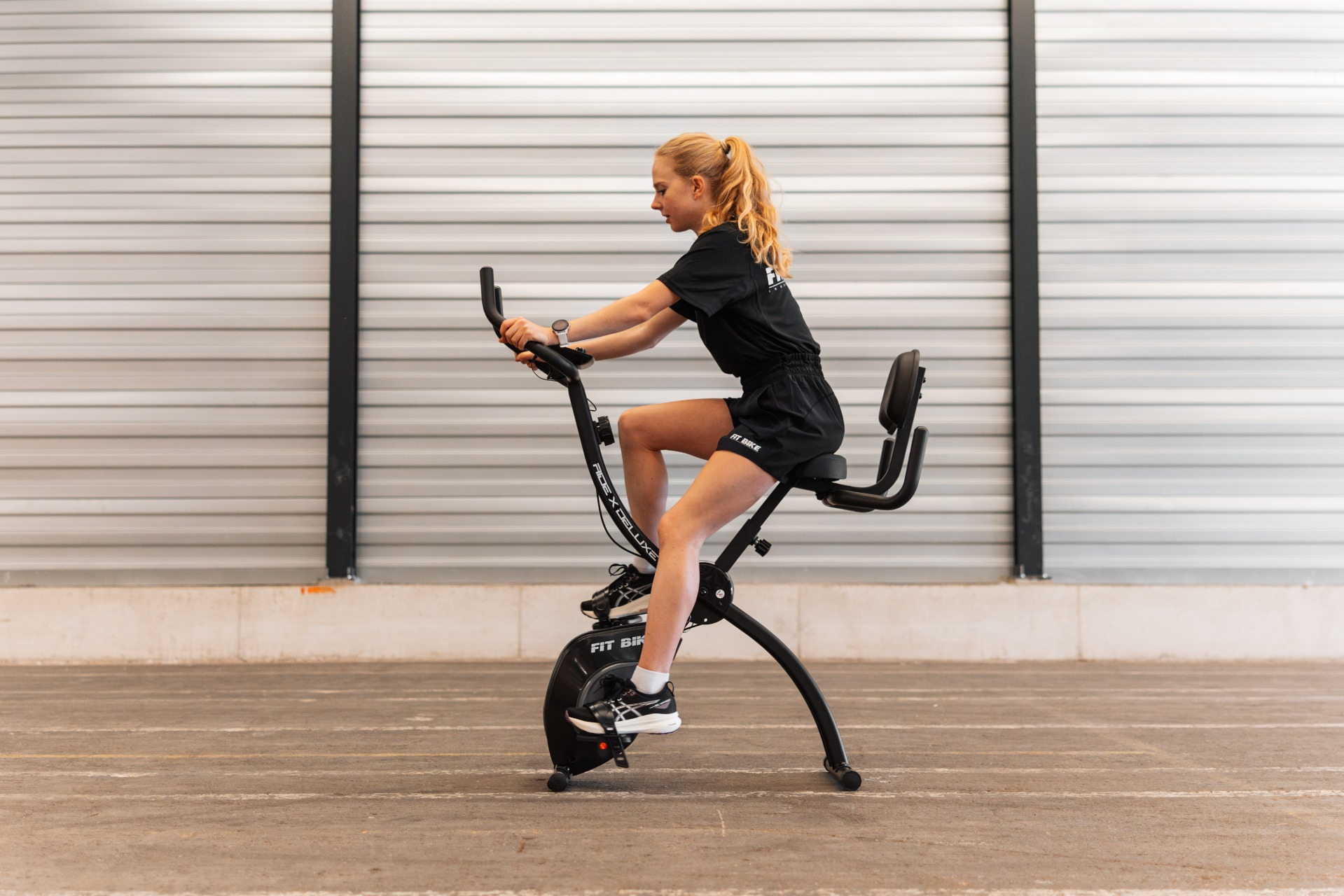 Een vrouw sport op een hometrainer. Ze draagt een zwarte korte broek en een shirt. De achtergrond is een metalen muur.