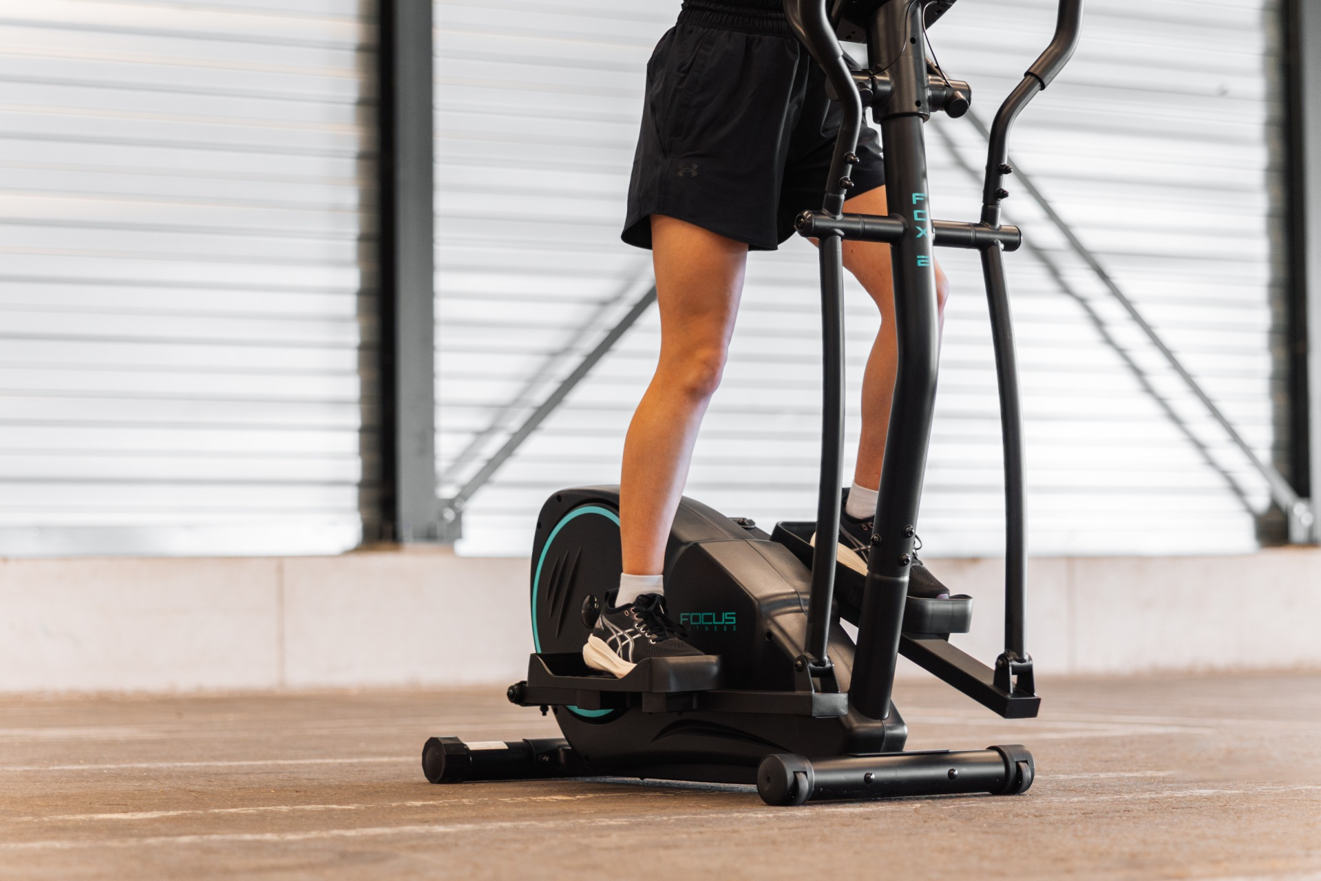 Une personne utilise un elliptique. Elle porte un short et des chaussures noirs. La machine est noire avec des accents bleus.