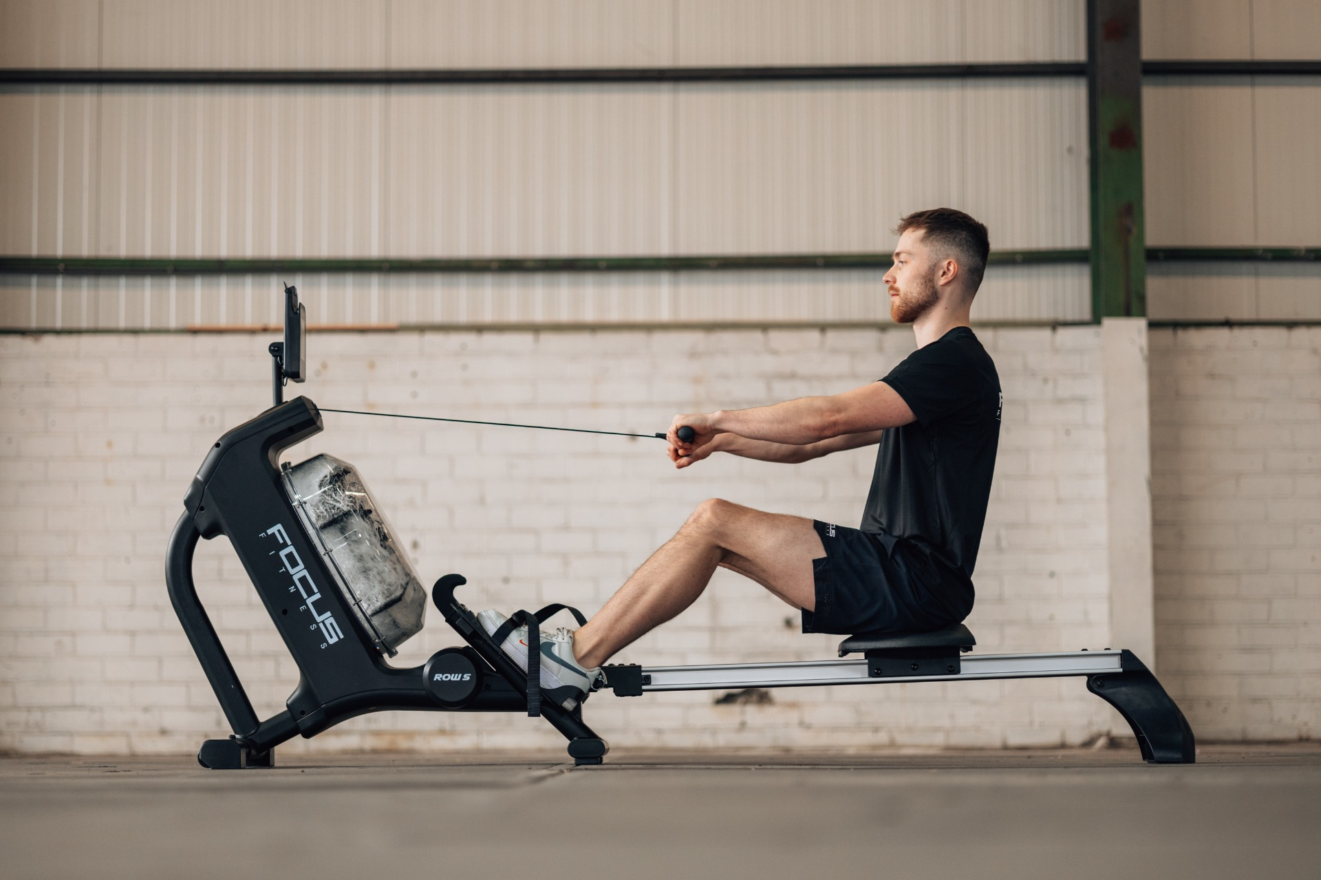 A man rows on a rowing machine. He wears black shorts and a shirt. The machine is black and has a water tank.