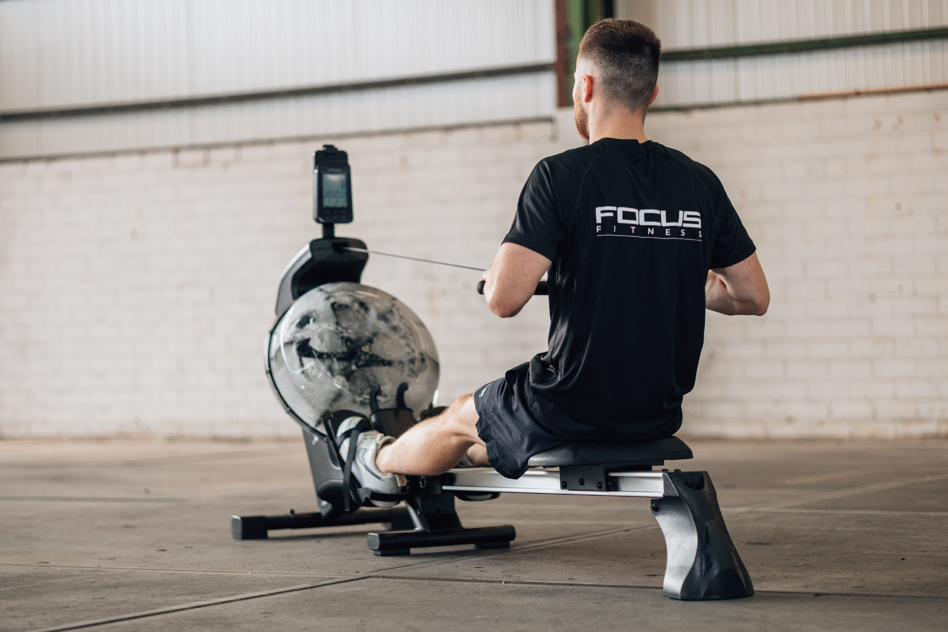 Man on rowing machine, back to camera. Black shirt, shorts. Focus Fitness logo. Gym setting, gray and white tones.