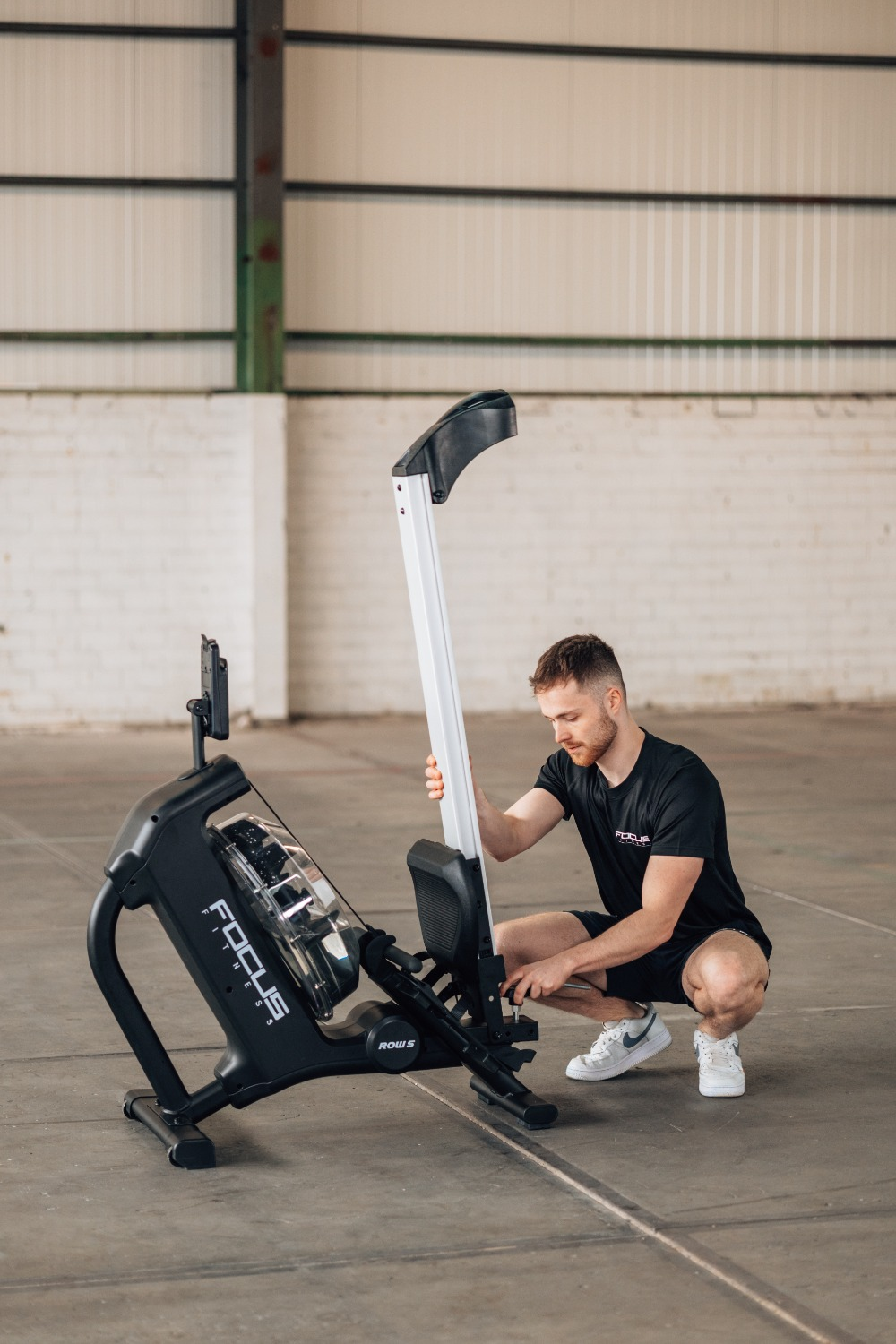 A man in a black shirt and shorts is assembling a black rowing machine in a warehouse setting.