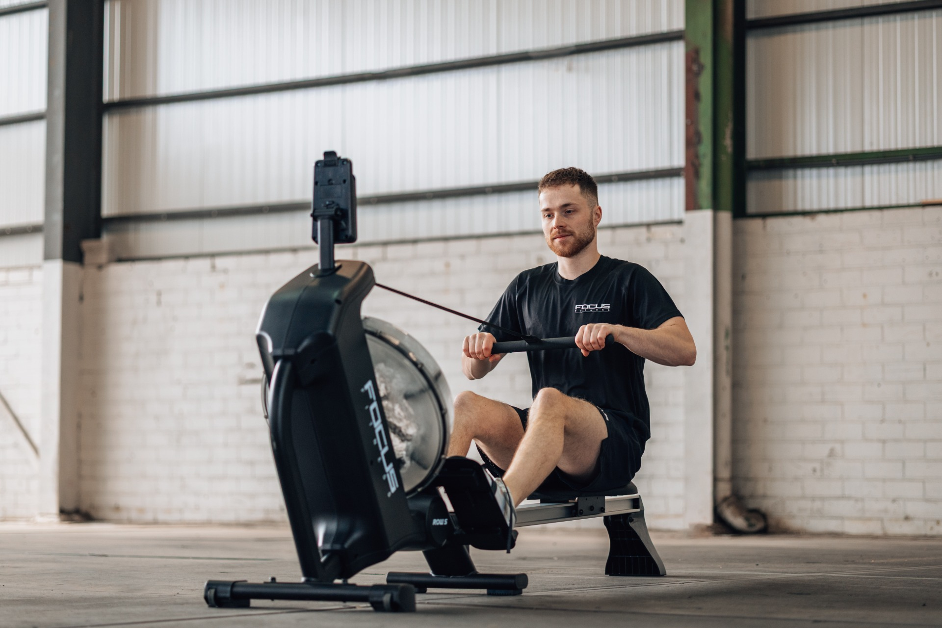 A man in black clothes using a rowing machine in a gym. The machine is black. Background is a white brick wall.