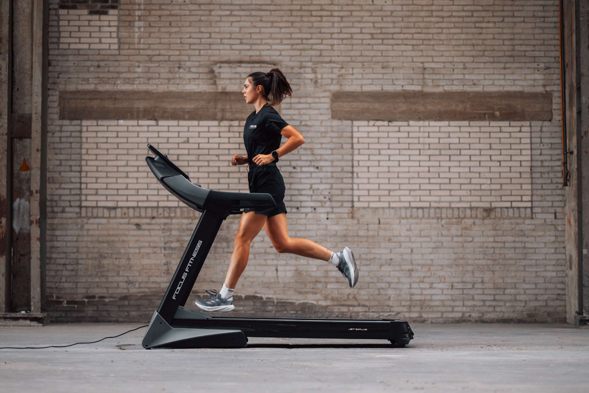 A woman runs on a treadmill. She wears black athletic attire. The background is a brick wall.