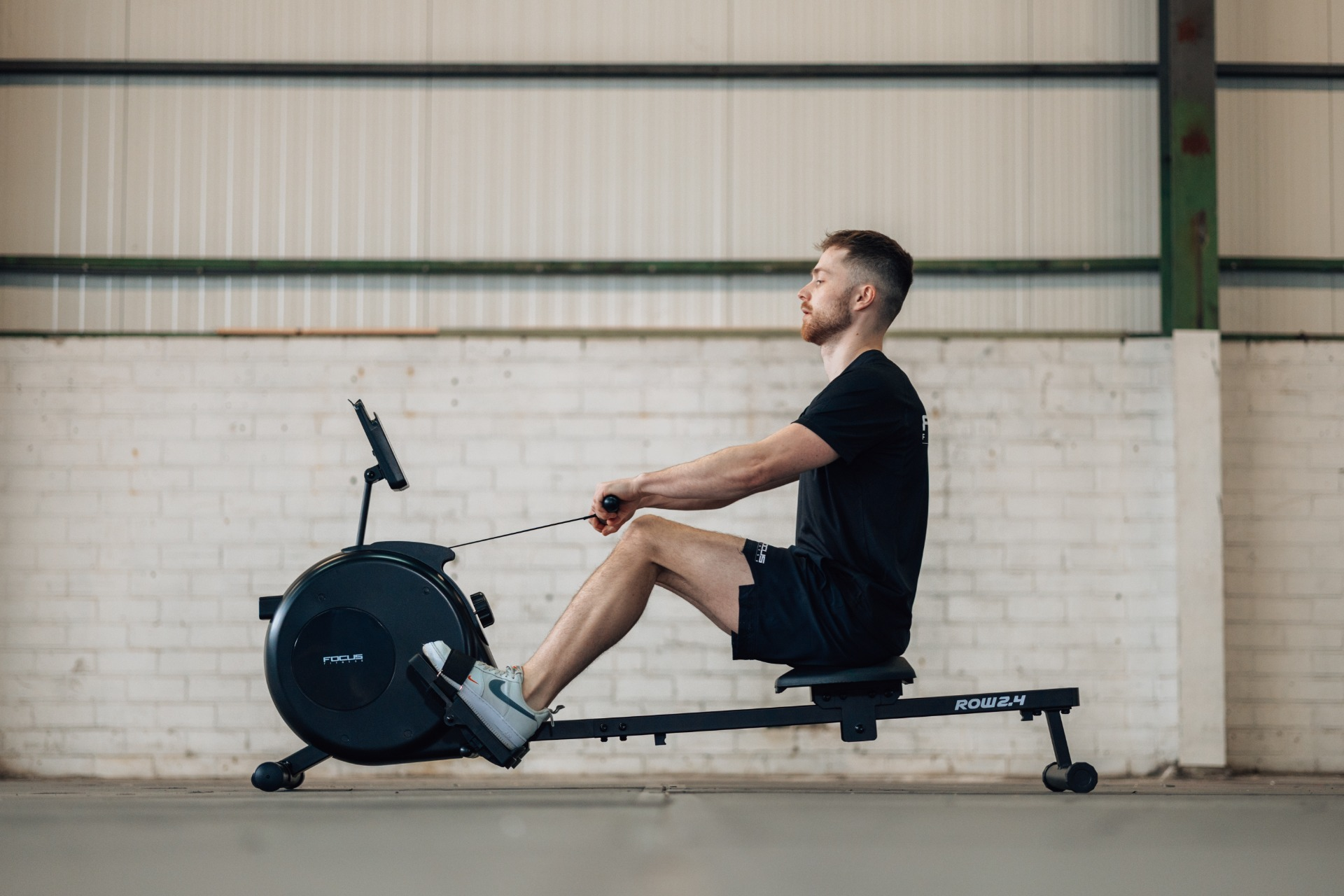 A man rows on a black rowing machine in a gym. He wears a black t-shirt and shorts. The background is a white brick wall.