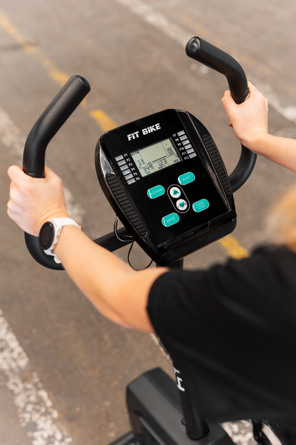 Une personne utilise un Fit Bike. Les mains tiennent le guidon. Une montre est au poignet gauche. L'écran affiche des chiffres et des boutons.