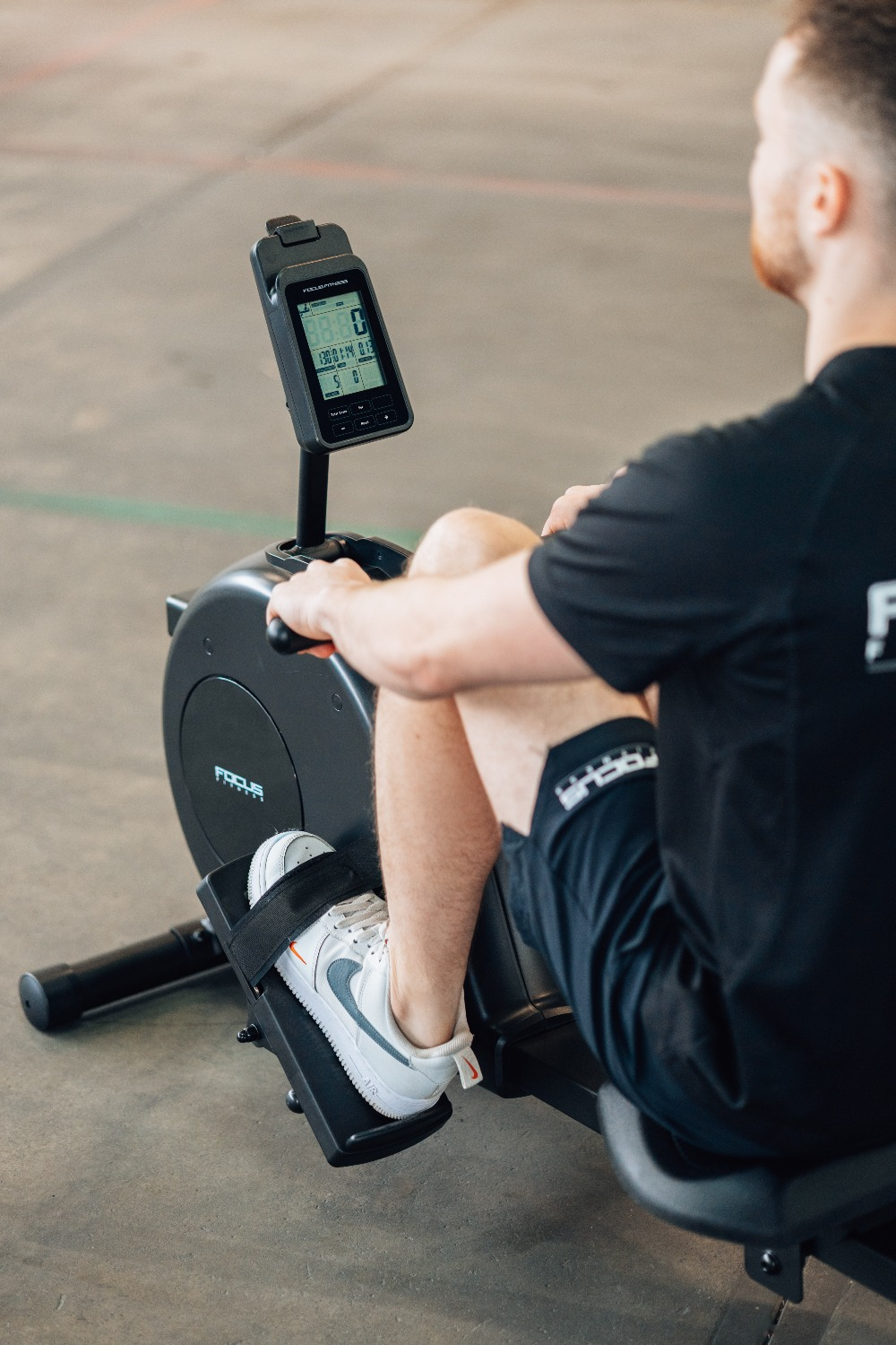 A man is using a Focus Fitness rowing machine. The display shows stats. He wears black shorts and a shirt, and white sneakers.
