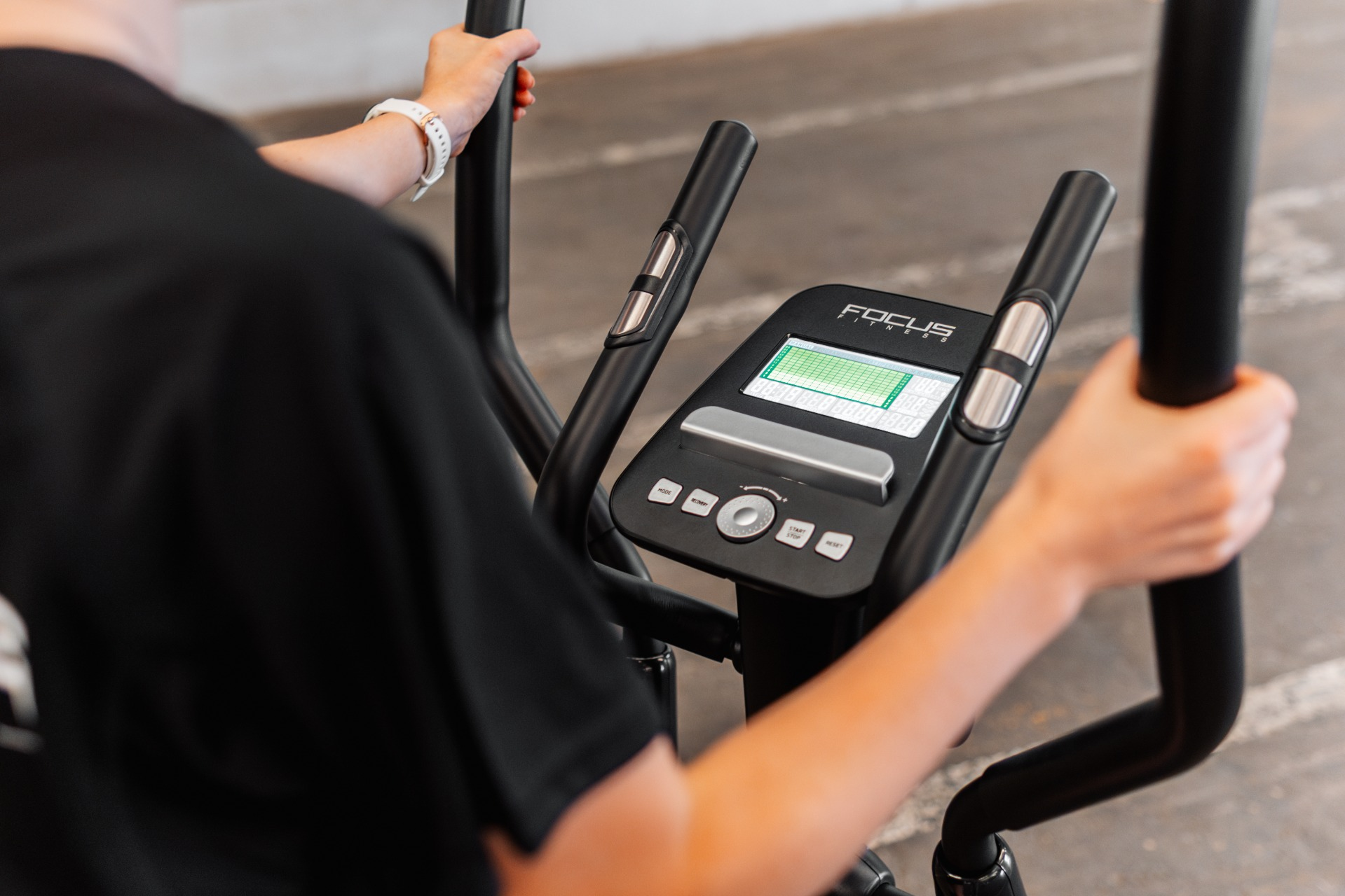 Une personne utilise un elliptique Focus Fitness. La machine a un écran numérique et des poignées noires. La personne porte une chemise noire.