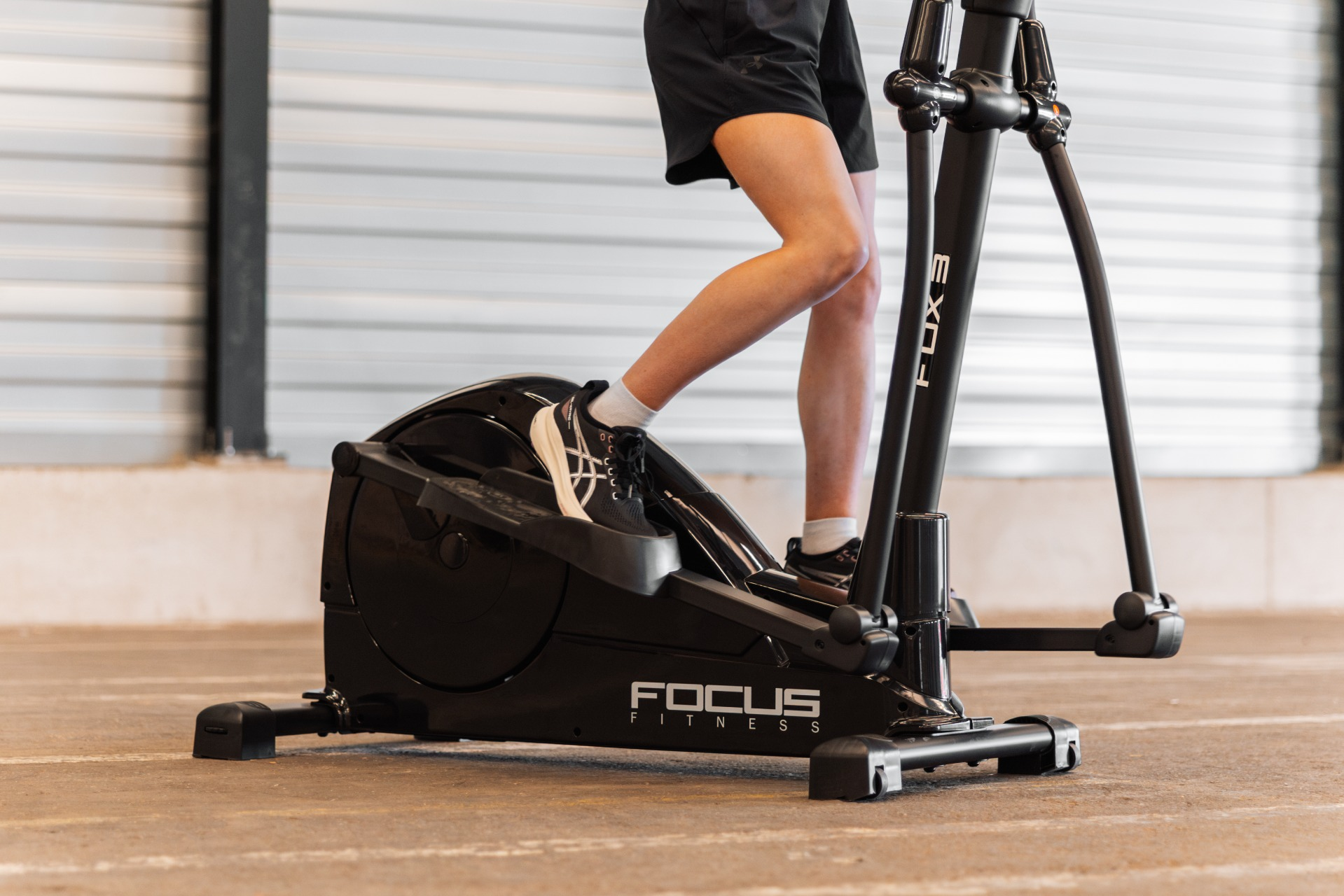 Une personne utilise un elliptique noir Focus Fitness. La personne porte un short et des chaussures. Le fond est un mur.