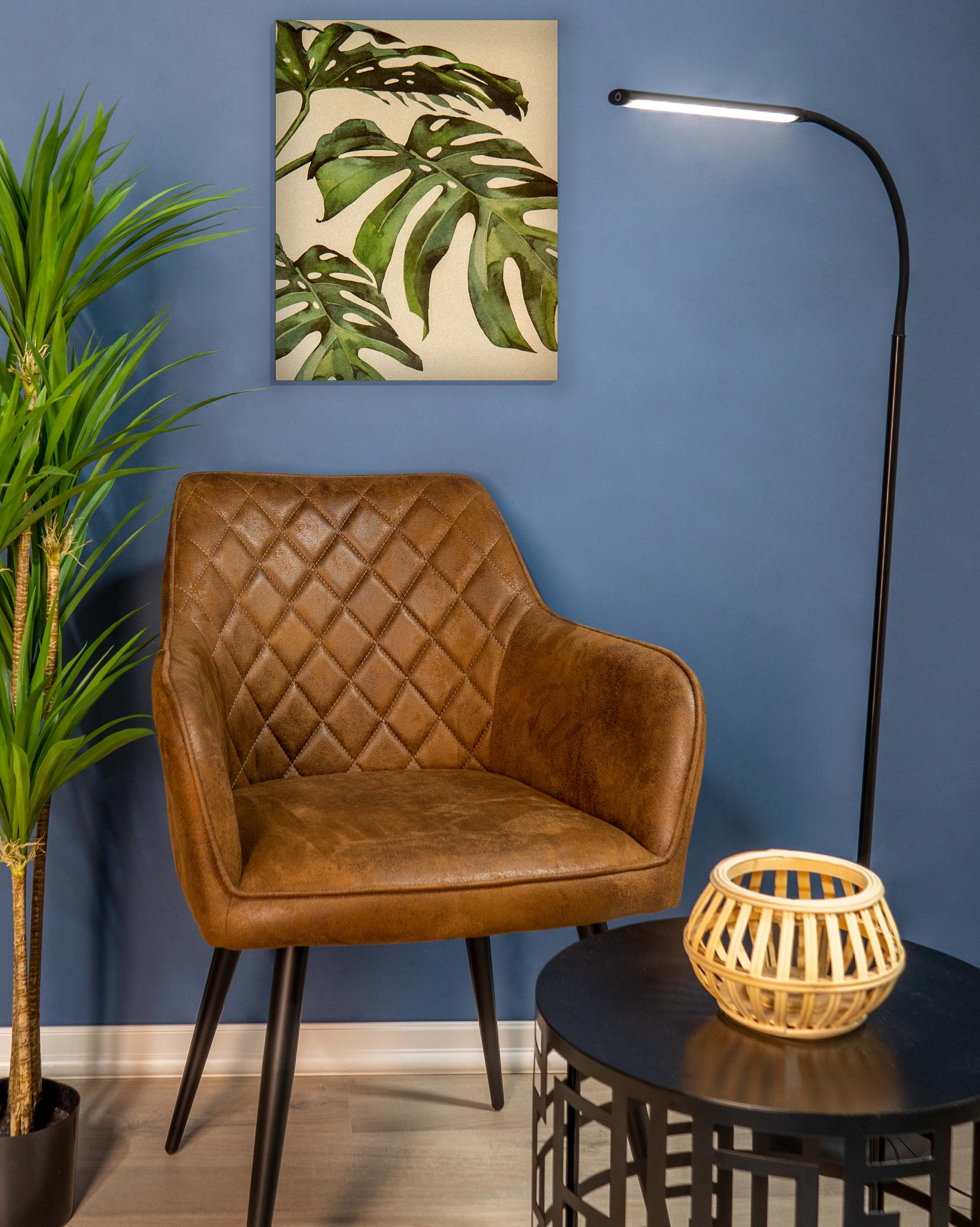 Una habitación con una silla marrón, planta, mesa auxiliar y un cuadro de hojas verdes. Pared azul.