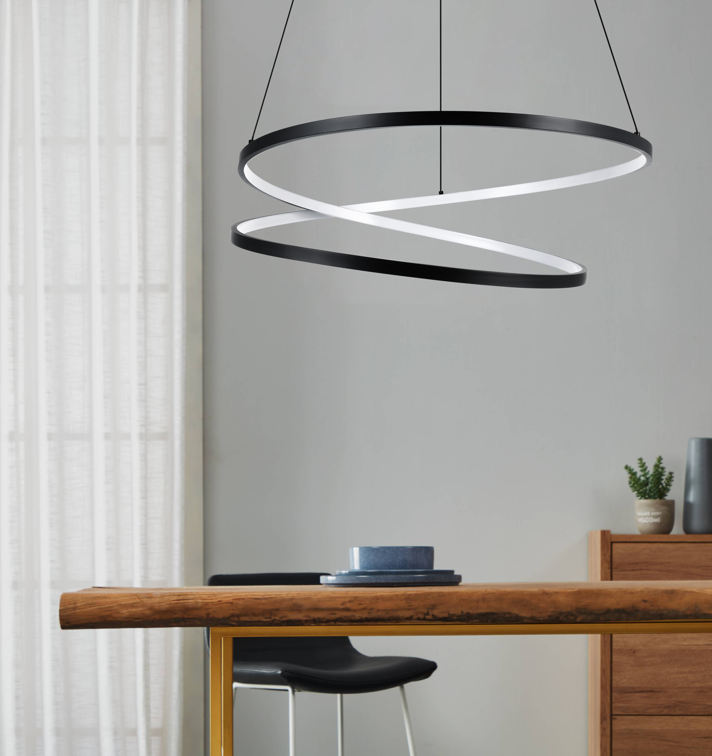Ein modernes Esszimmer. Über einem Holztisch hängt eine schwarze, kreisförmige Leuchte. Ein Fenster mit weißen Vorhängen.
