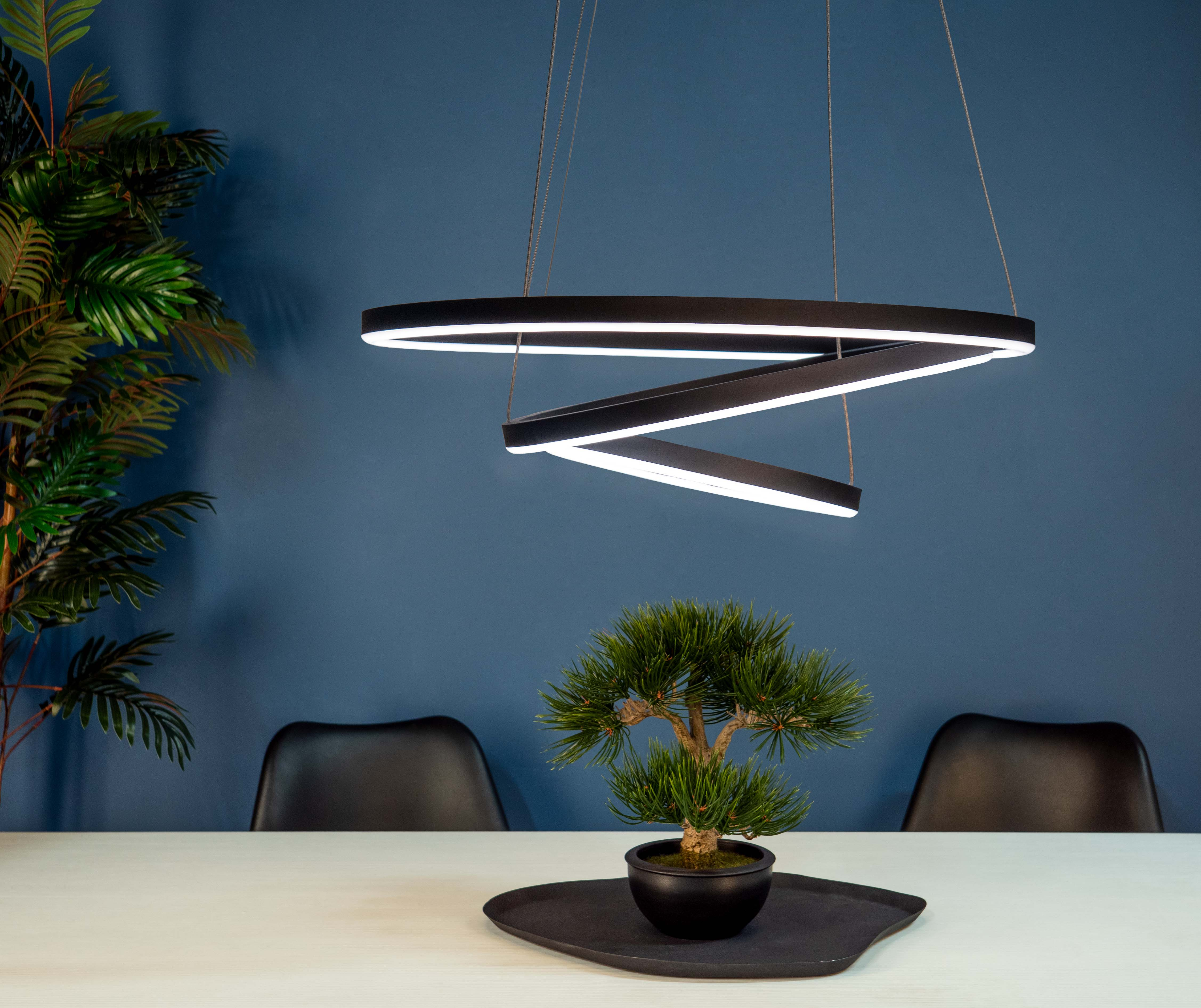 Eine moderne schwarze Pendelleuchte hängt über einem Tisch mit einem Bonsai-Baum. Tiefblaue Wand im Hintergrund.