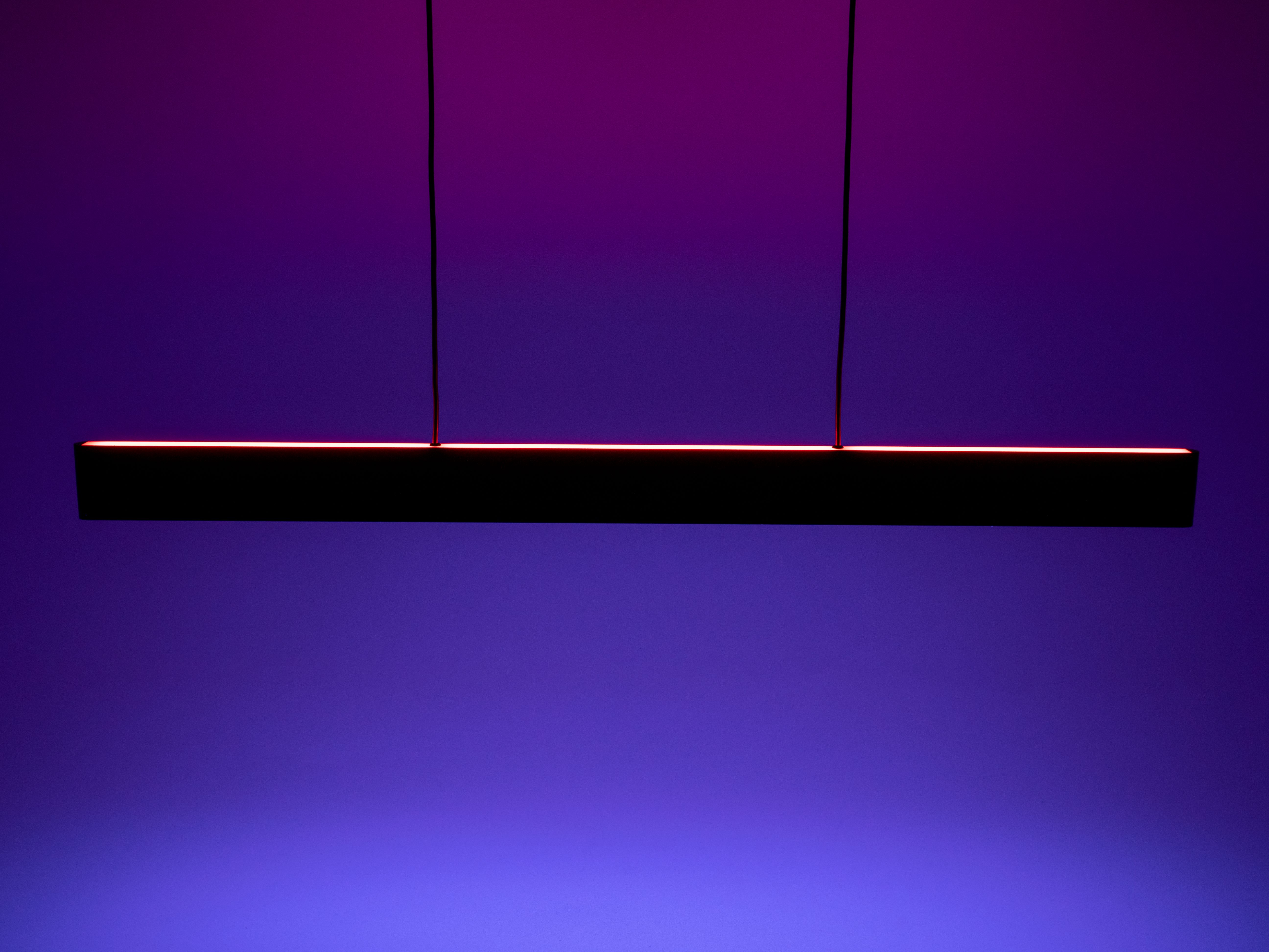 Un long luminaire noir est suspendu à deux fils sur un fond dégradé violet. La lumière est allumée.